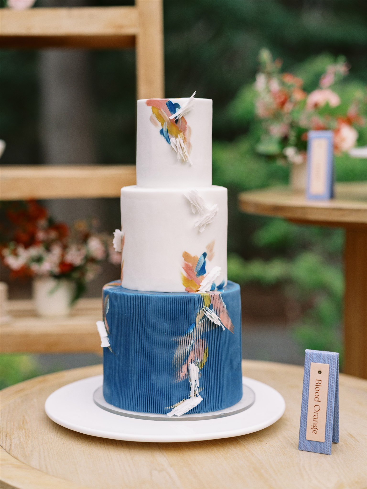 A tiered cake decorated with white, blue, and multicolored abstract design elements, displayed on a wooden table with a pink and cream ribbon beside it.