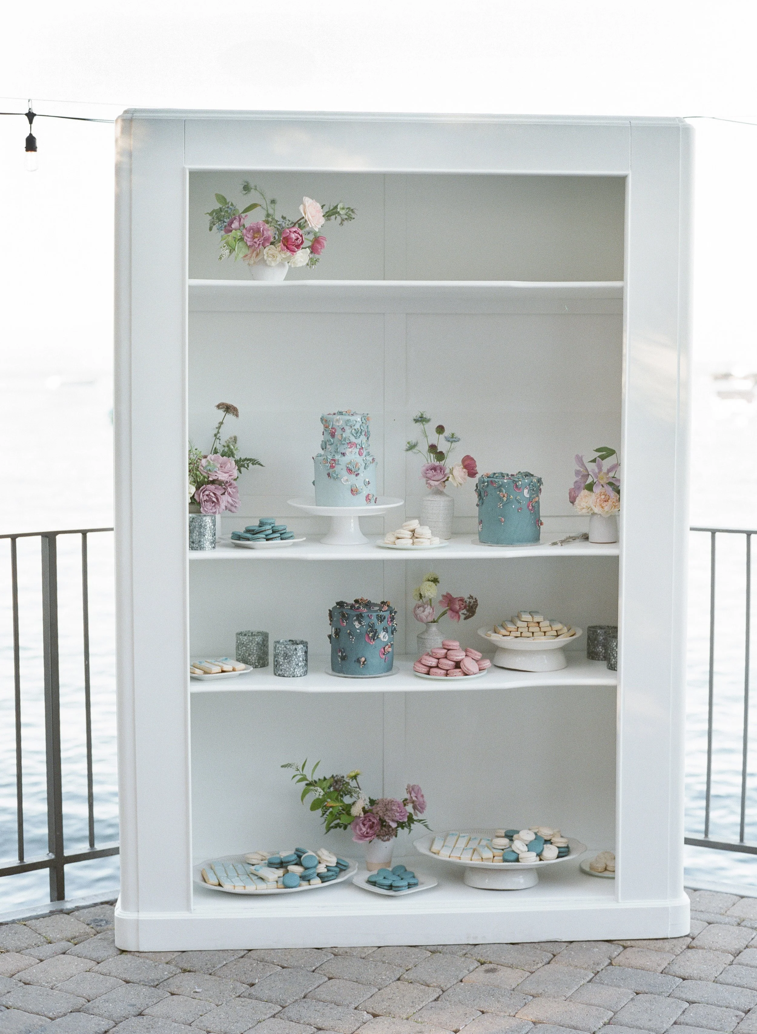 A white display shelf with flowers, decorated cakes, and baked goods, outdoors by a body of water, with a string of outdoor lights above.