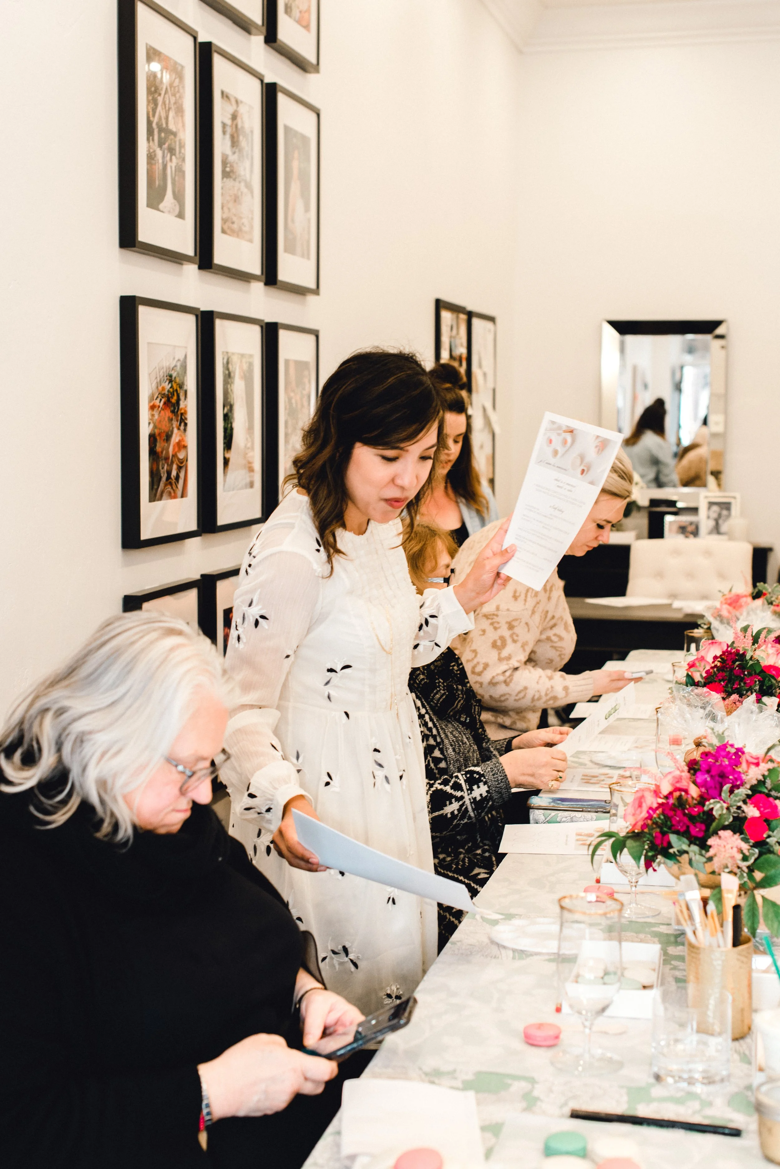 Women gathered around a table with floral arrangements, looking at menus or papers in an elegant indoor setting decorated with artwork on the wall.