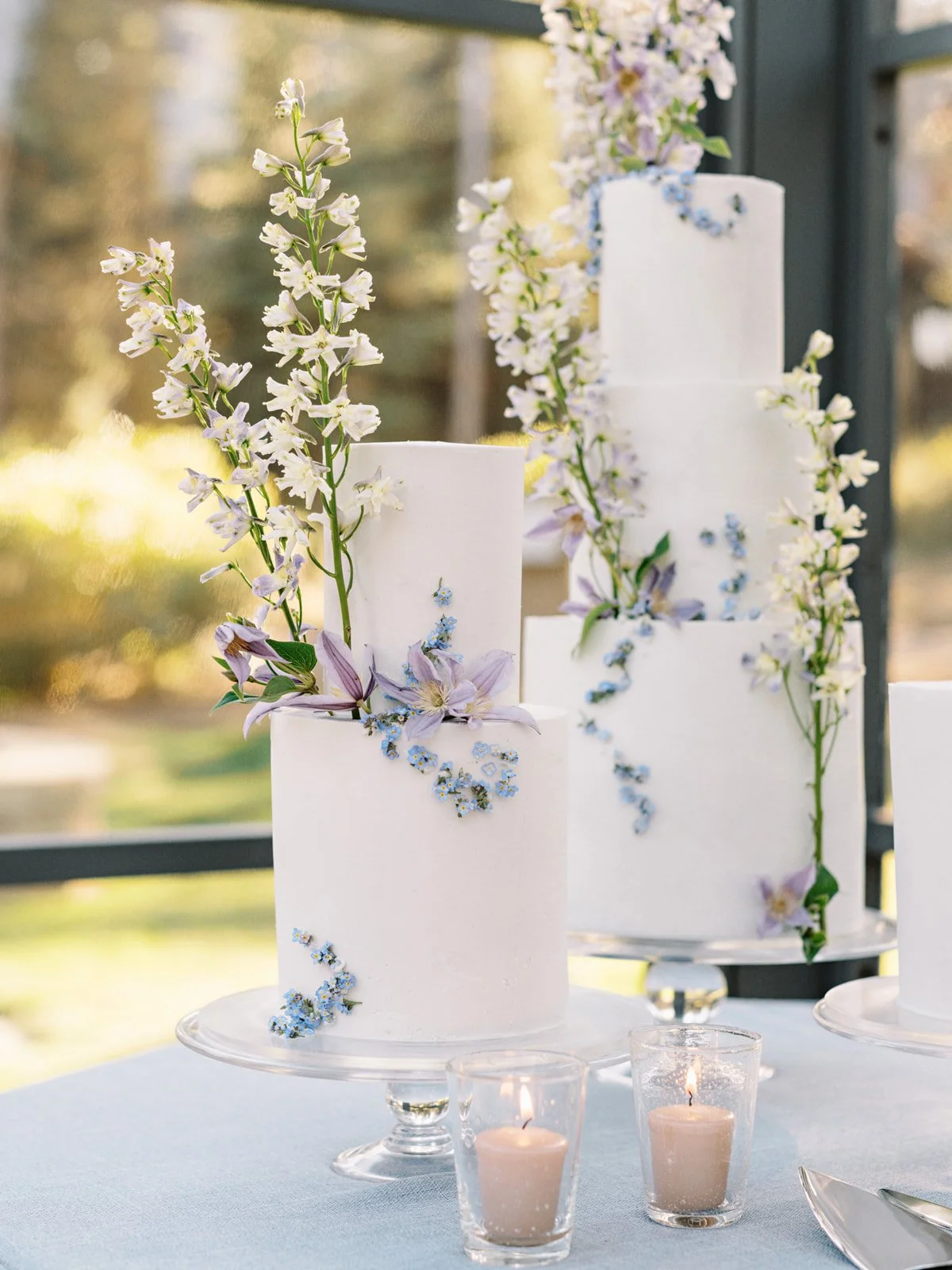 Elegant white wedding cake decorated with fresh flowers including lavender, lilies, and small blue and white blossoms, placed on a white table with candles.