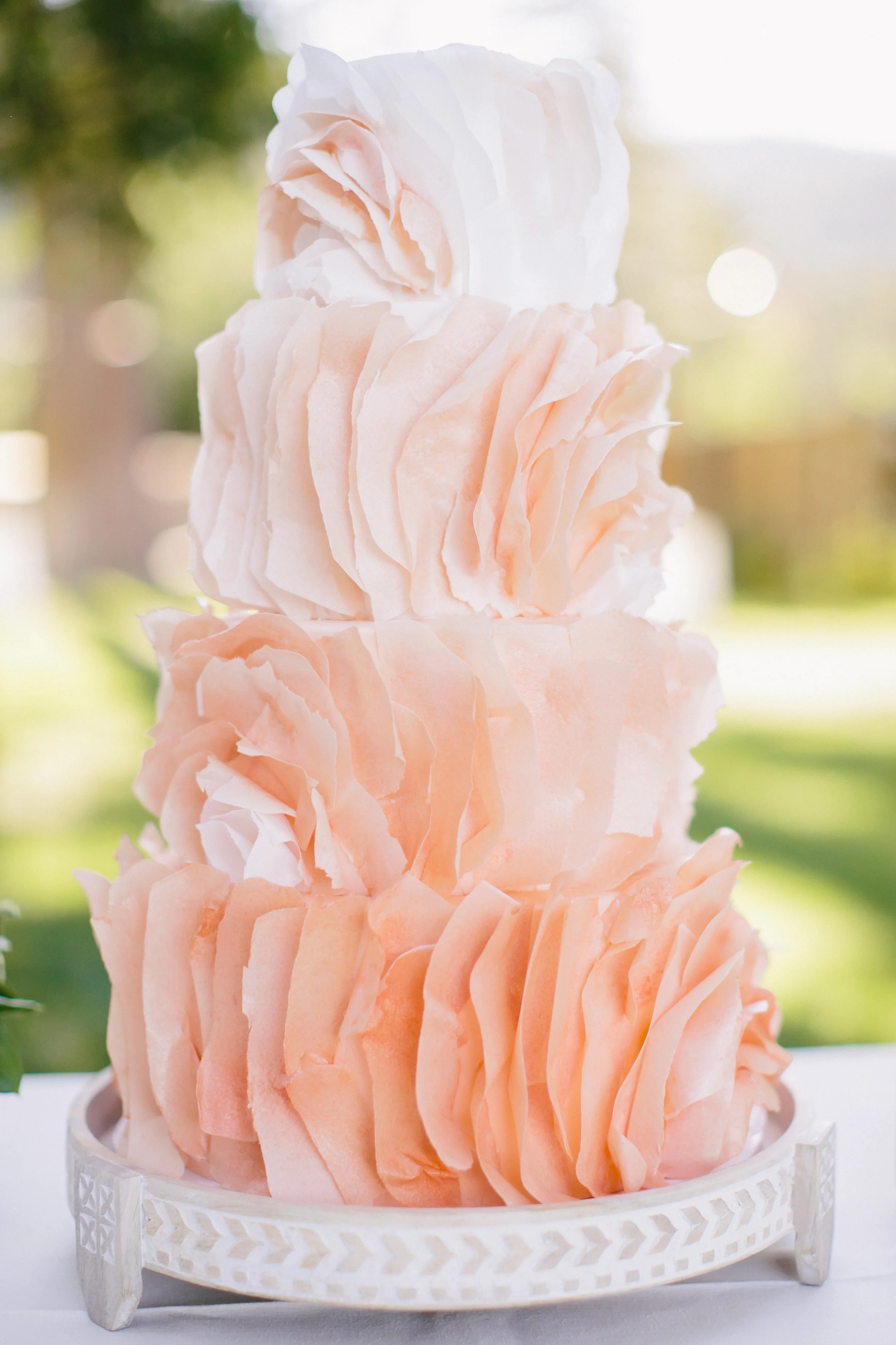 A two-tiered cake decorated with light pink and white ruffled buttercream frosting, placed on a white decorative stand with a geometric pattern. The cake is outdoors with a blurred green and light background.
