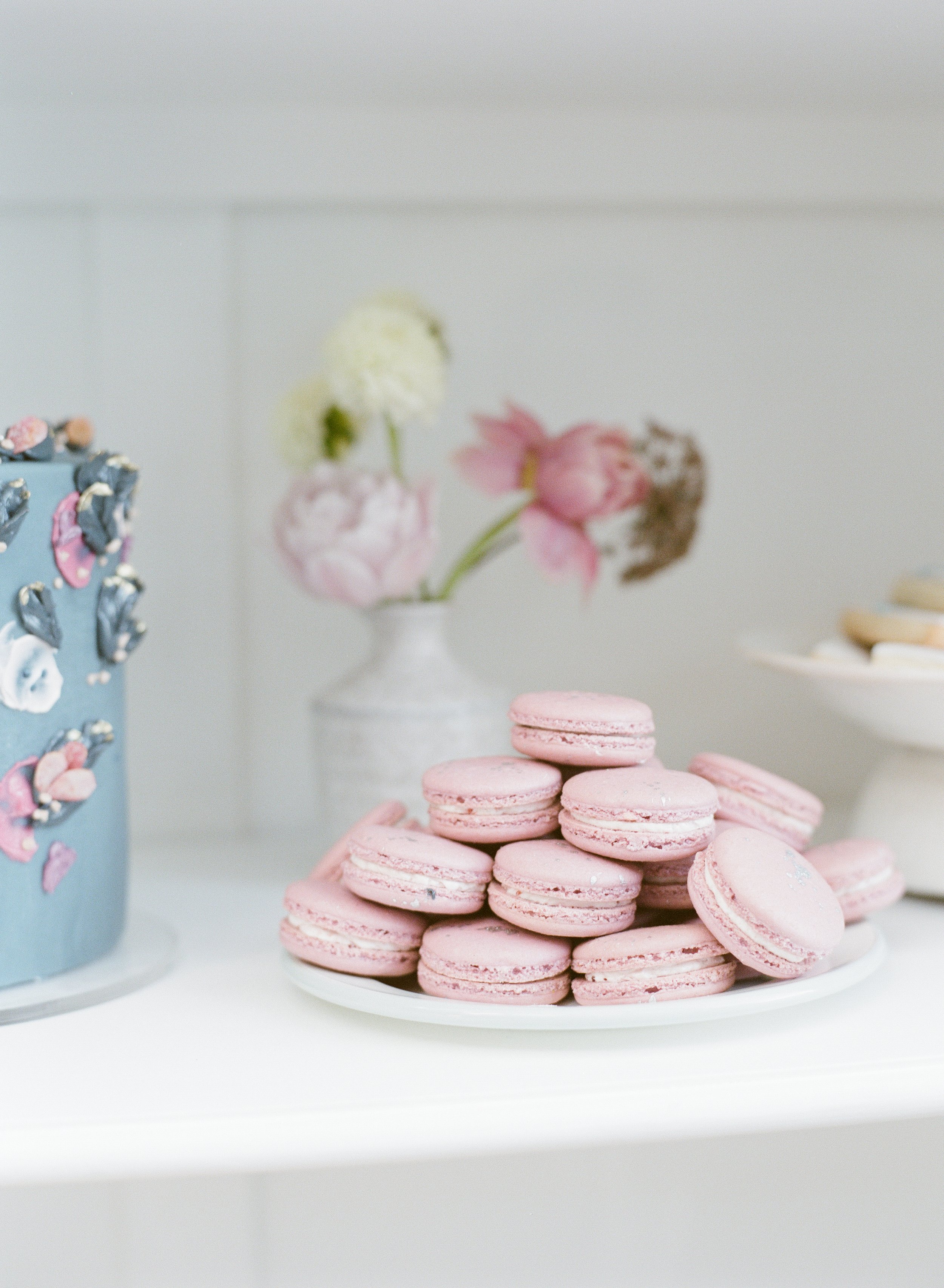 A white plate with pink macarons stacked on top, in the background is a vase with pink and white flowers, and a partially visible decorated cake on the left.