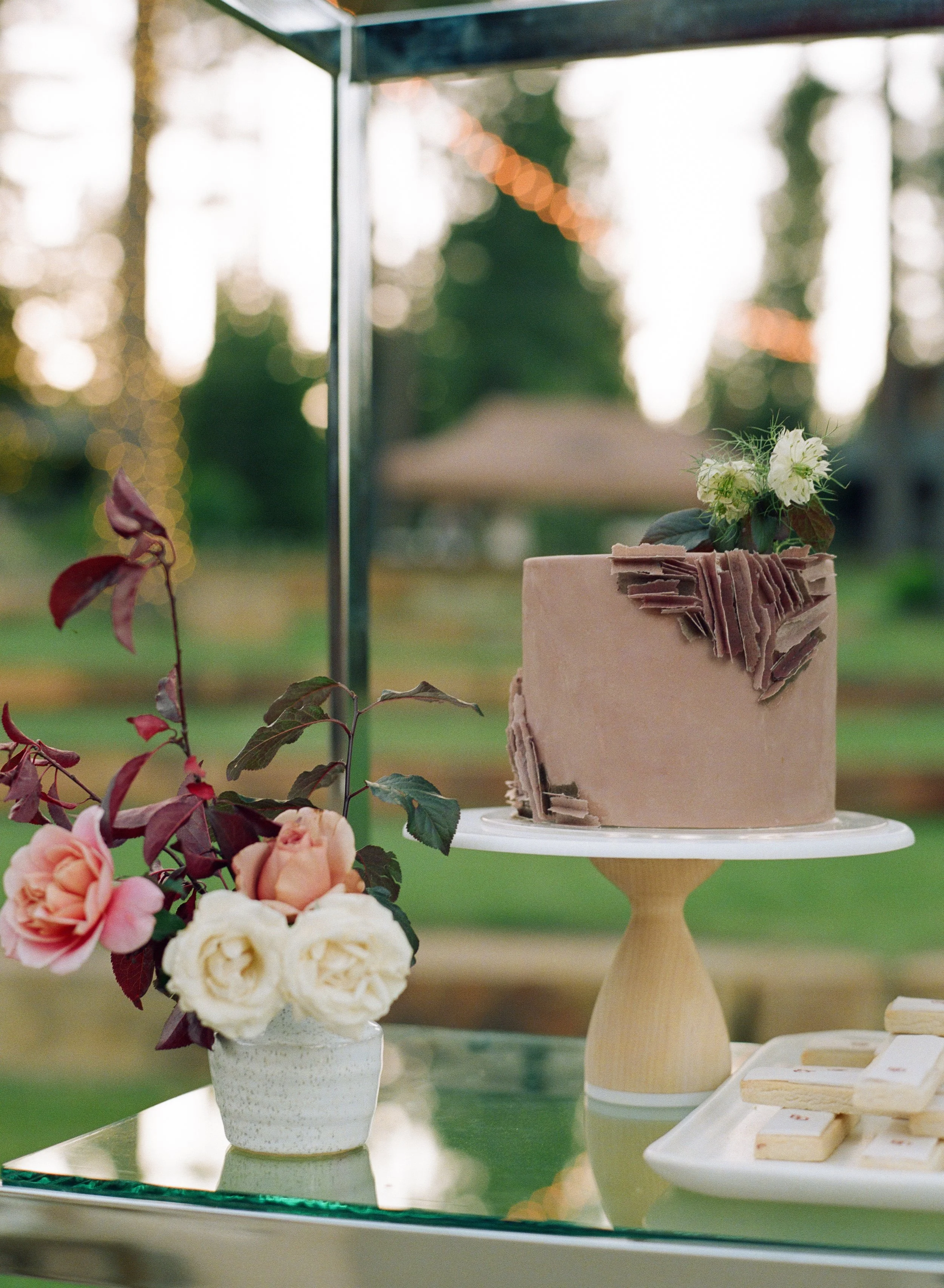 A bakery display featuring a pink cake with flower decorations, surrounded by flower arrangements, on a reflective surface outdoors with trees in the background.