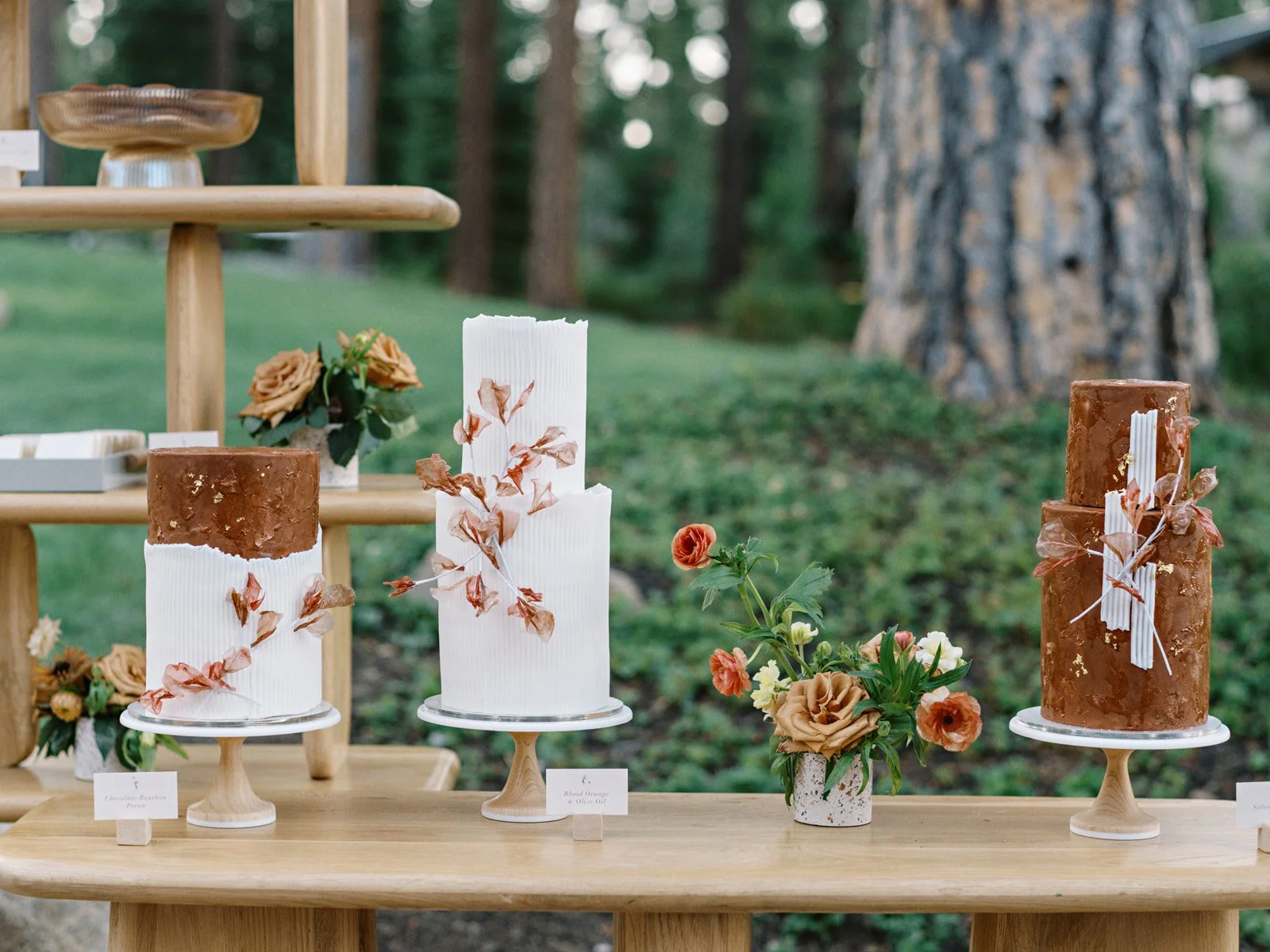 Three tall cylindrical cakes with brown frosting and decorative dried leaves, displayed on white stands outdoors with flowers and greenery in the background.