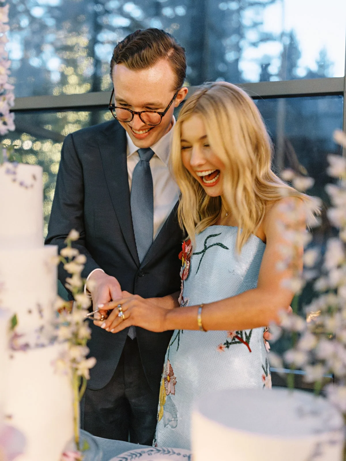 A smiling couple, a man in a suit and glasses and a woman in a floral dress, are happily cutting a wedding cake together.