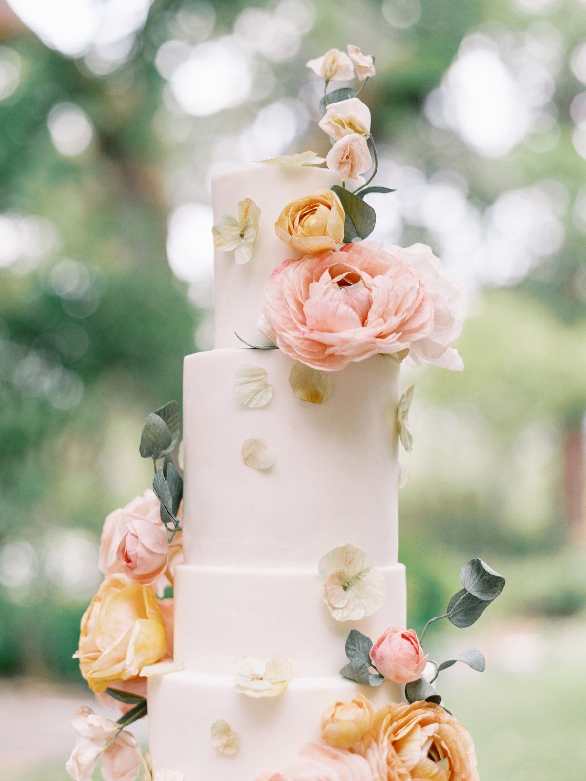 Elegant three-tier white wedding cake decorated with pink and yellow roses, green leaves, and scattered petals, set against a blurred outdoor background.