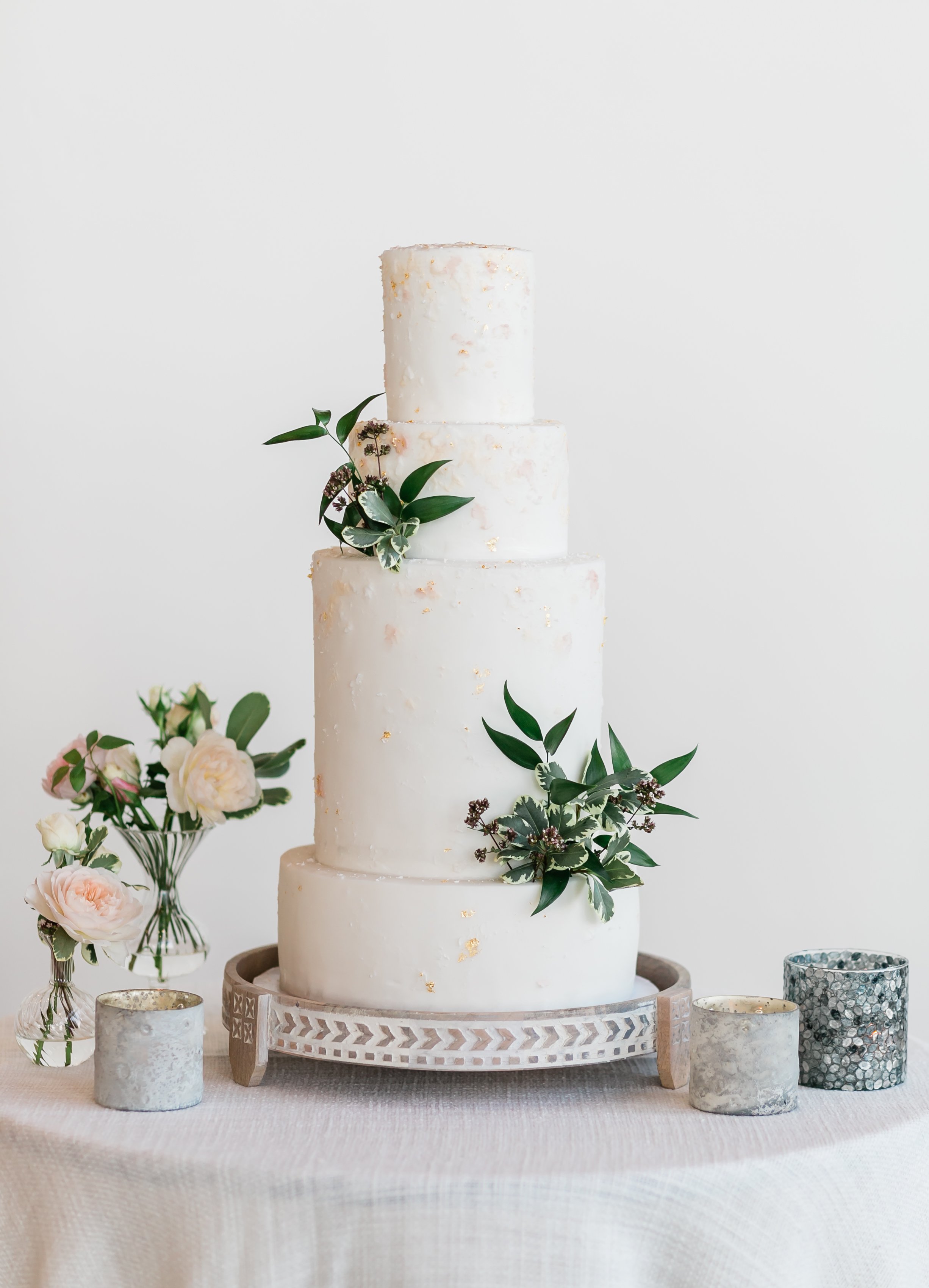A tiered white wedding cake decorated with green leaves and gold accents, set on a patterned stand. Surrounding the cake are candles in silver and marble holders, with pink and white flowers in a vase nearby.