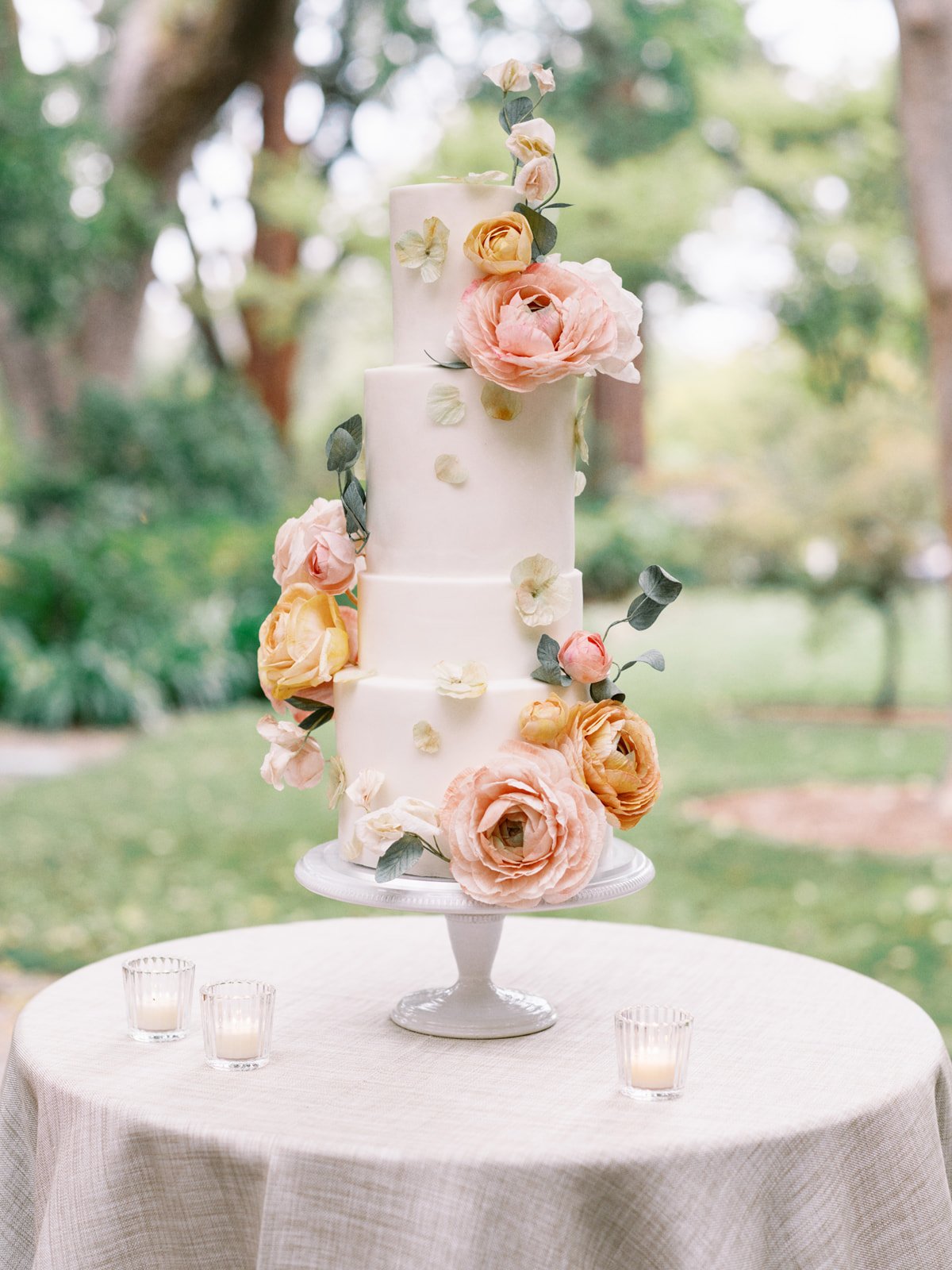 A tall, white three-tiered wedding cake decorated with pink and peach flowers and green leaves, placed on a white cake stand outdoors on a round table with white tablecloth. Small candles are arranged around the cake on the table, and trees and green