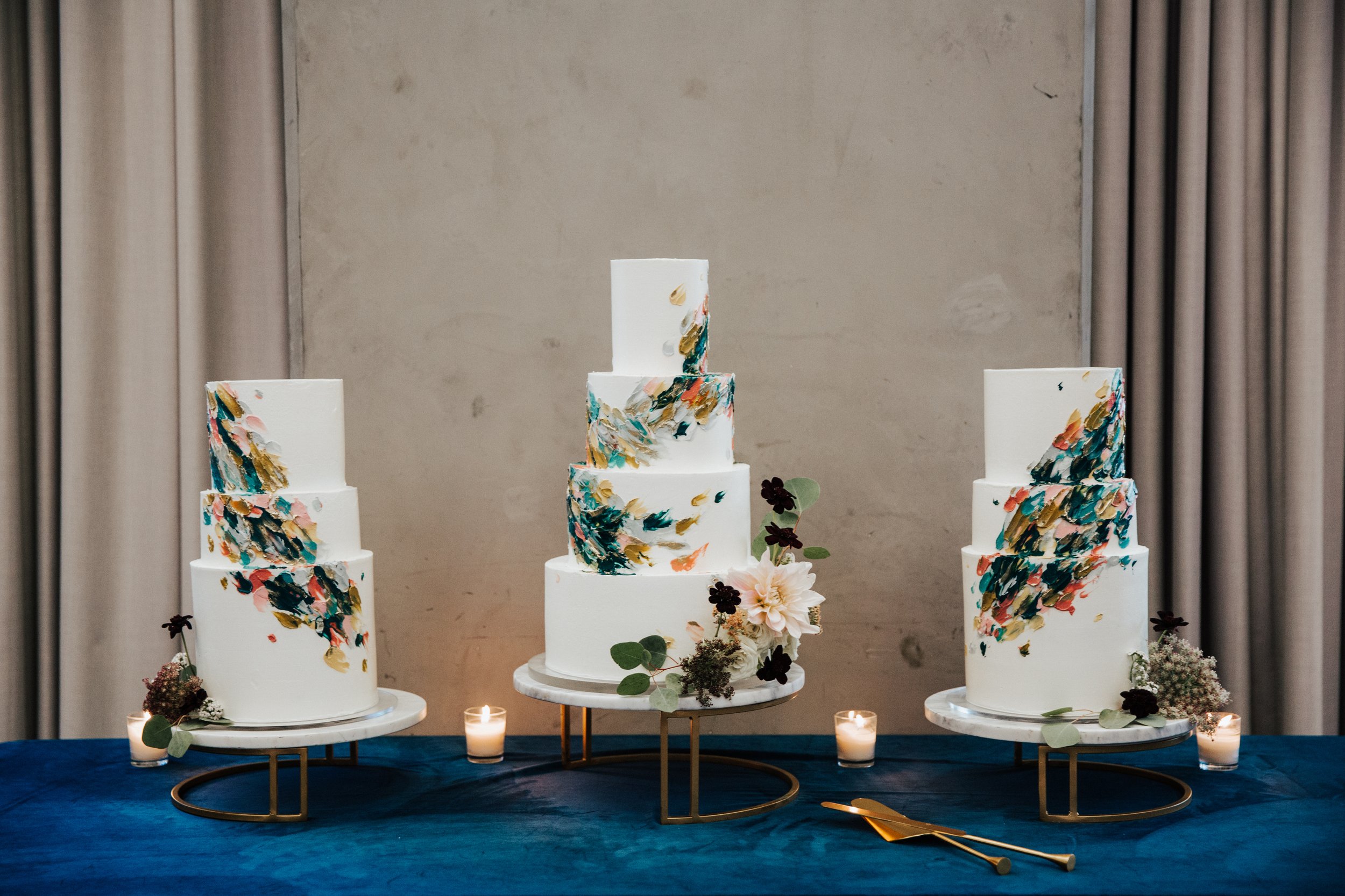 A multi-tiered white wedding cake with colorful abstract brushstroke designs, decorated with flowers and greenery, displayed on tables with candles around it.