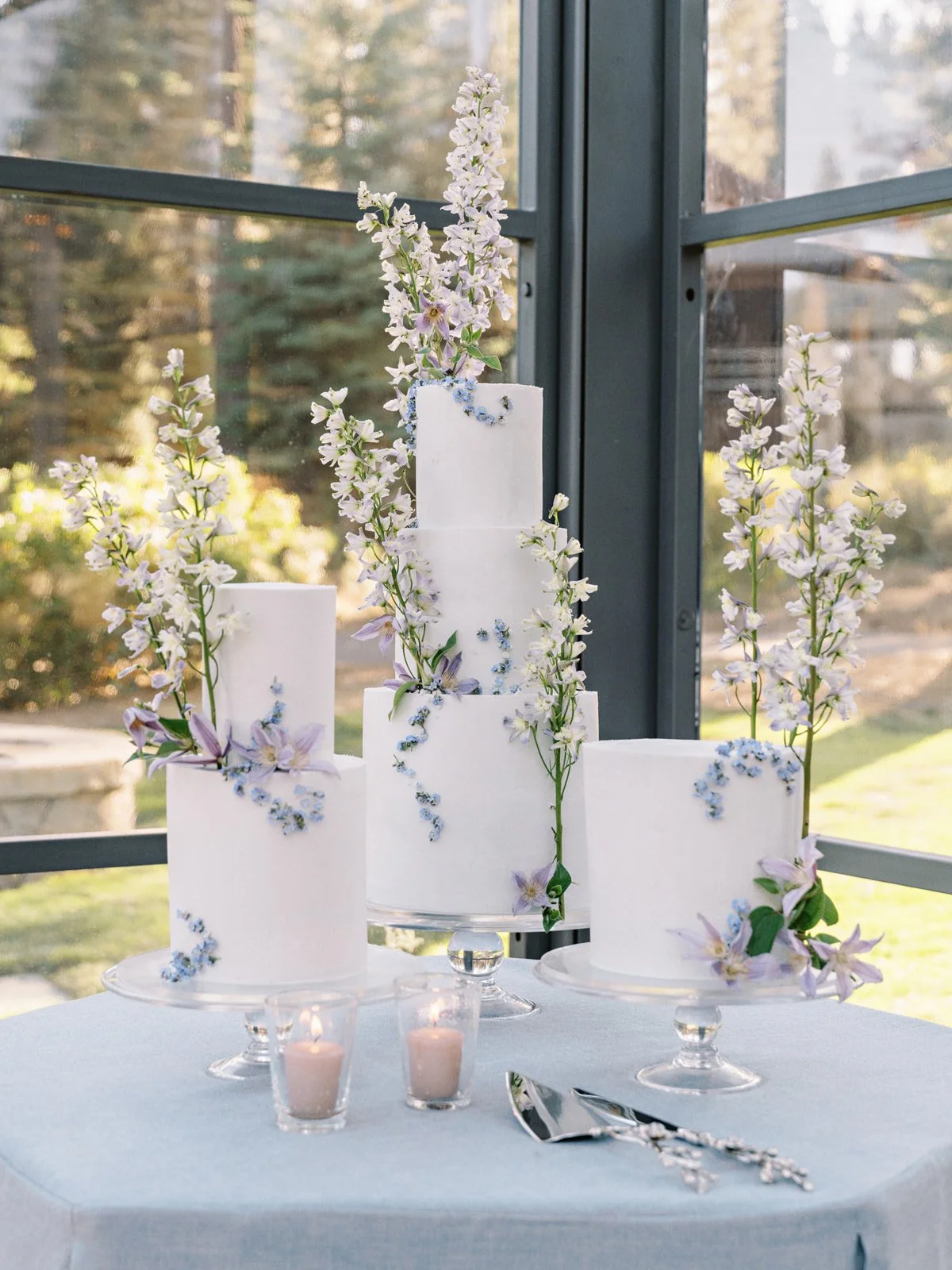 A multi-tiered white wedding cake decorated with pastel flowers, set on a table with small candles and silver cake server, with a glass window and outdoor scenery in the background.