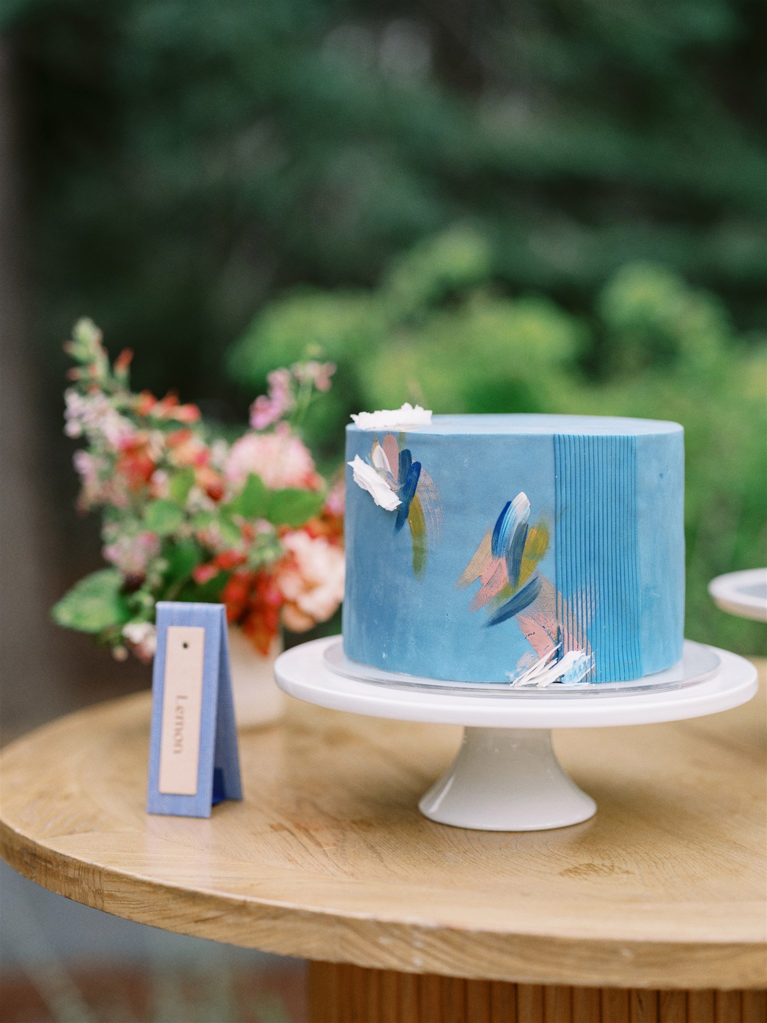 A small cake with blue and abstract painted design on a white cake stand, small flowers in the background, on a wooden table.
