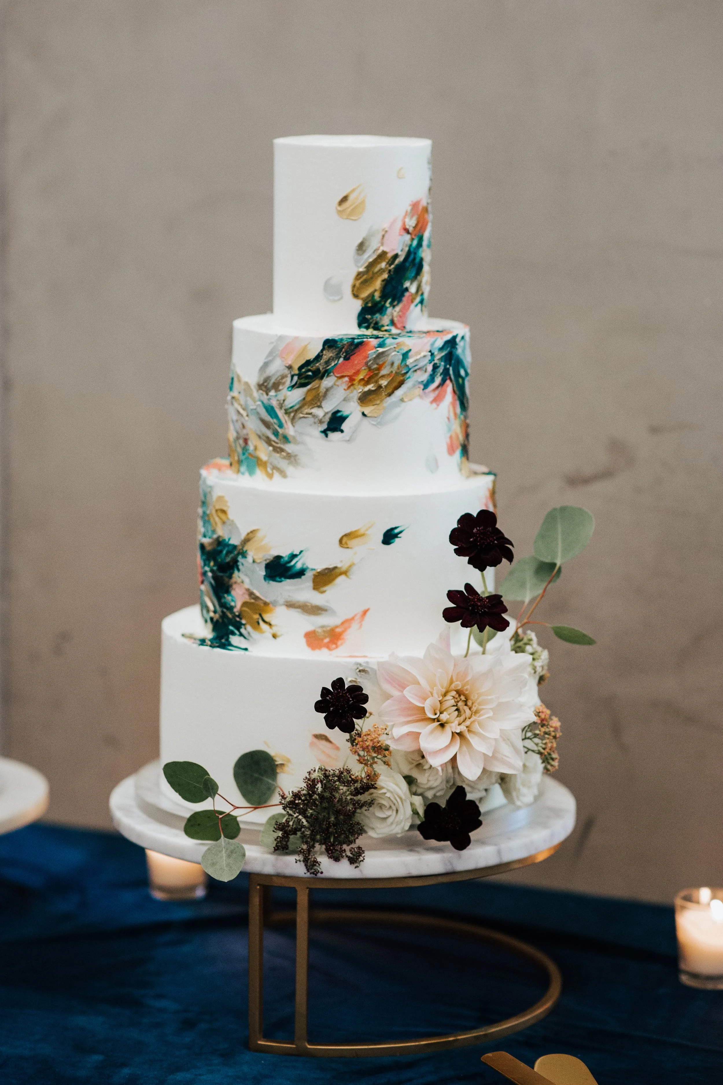 A three-tiered white wedding cake with colorful abstract brushstroke patterns and floral decorations, placed on a marble cake stand with a gold circular base, against a neutral background.