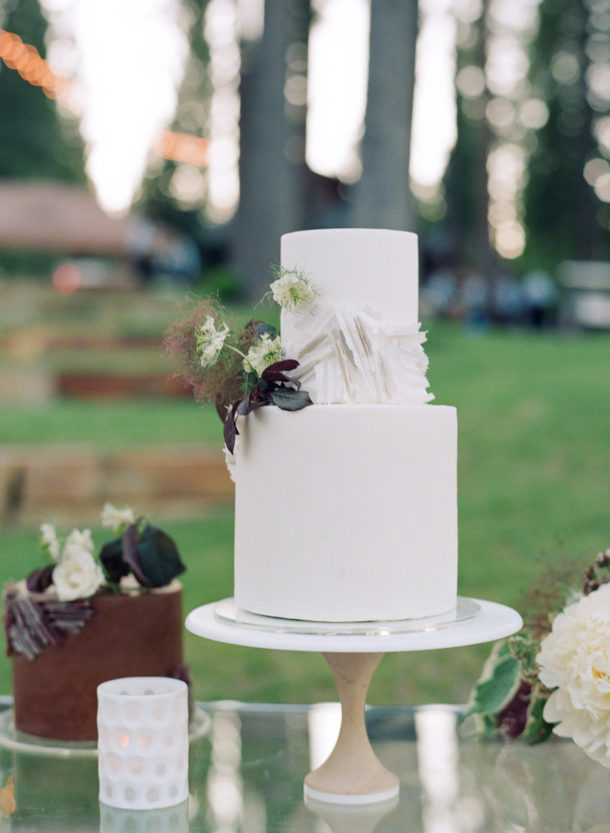 A white tiered cake decorated with white flowers and dark leaves, displayed outdoors on a stand with trees and blurred lights in the background.