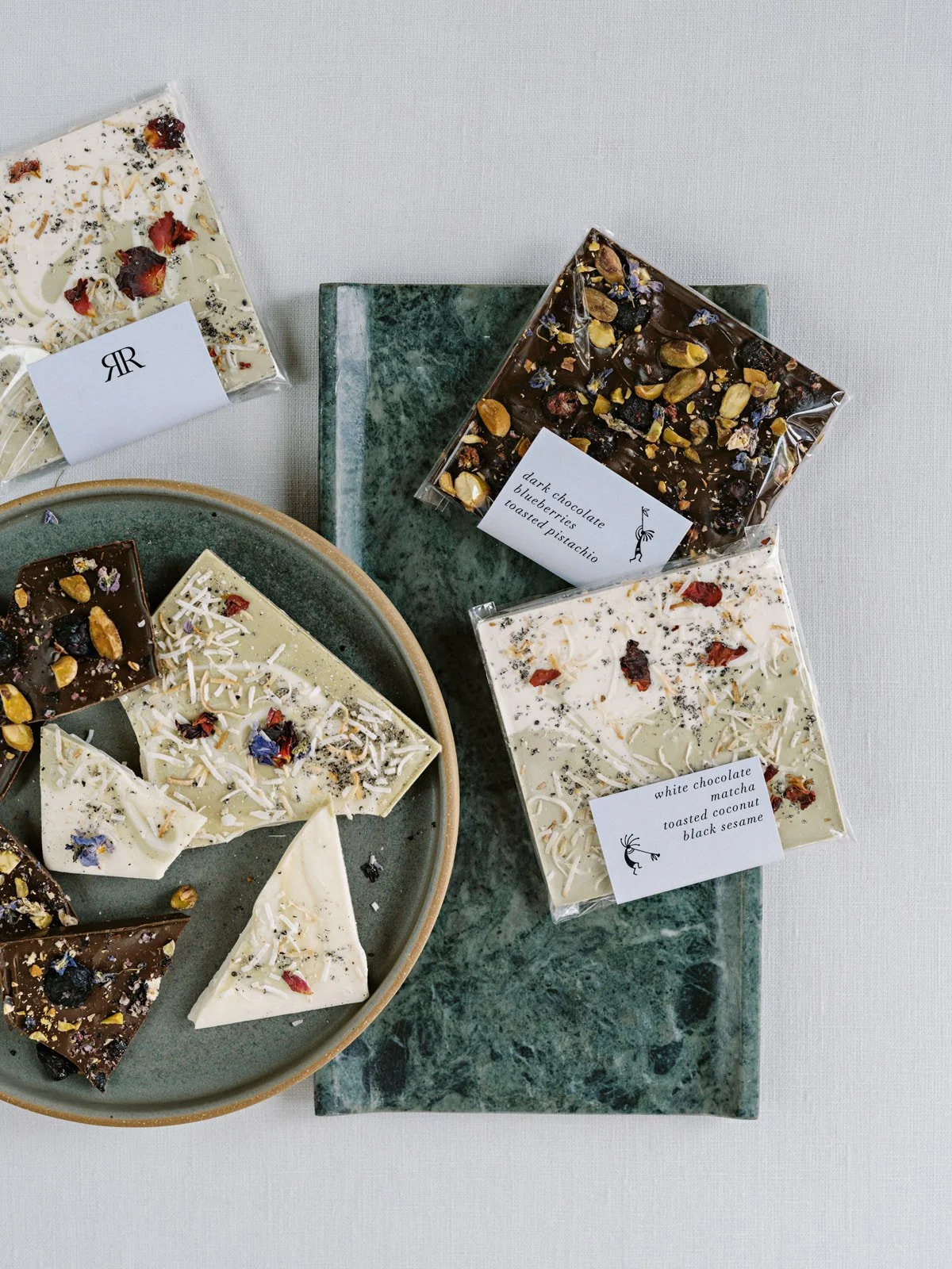 Assorted white and dark chocolates topped with nuts, dried berries, and edible flowers, some in clear plastic packaging with labels indicating flavors.