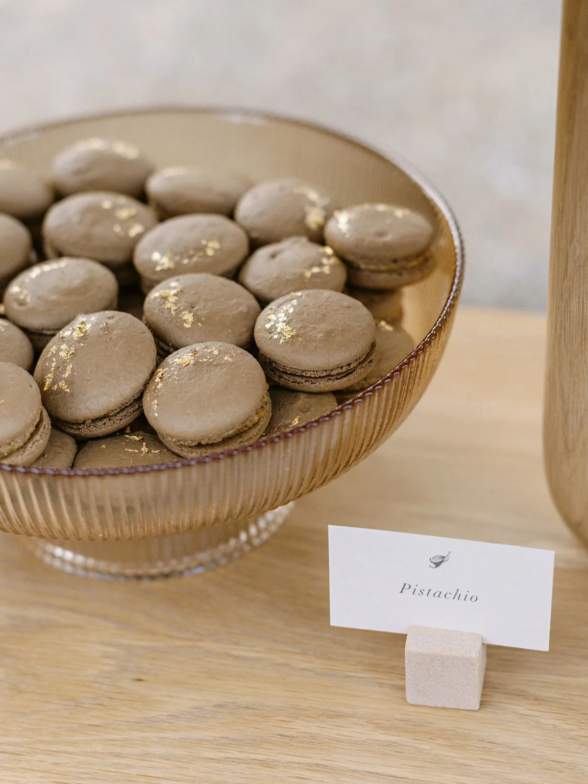 A bowl of pistachio-flavored macarons with gold flakes, displayed on a wooden surface with a small label that reads 'Pistachio'.