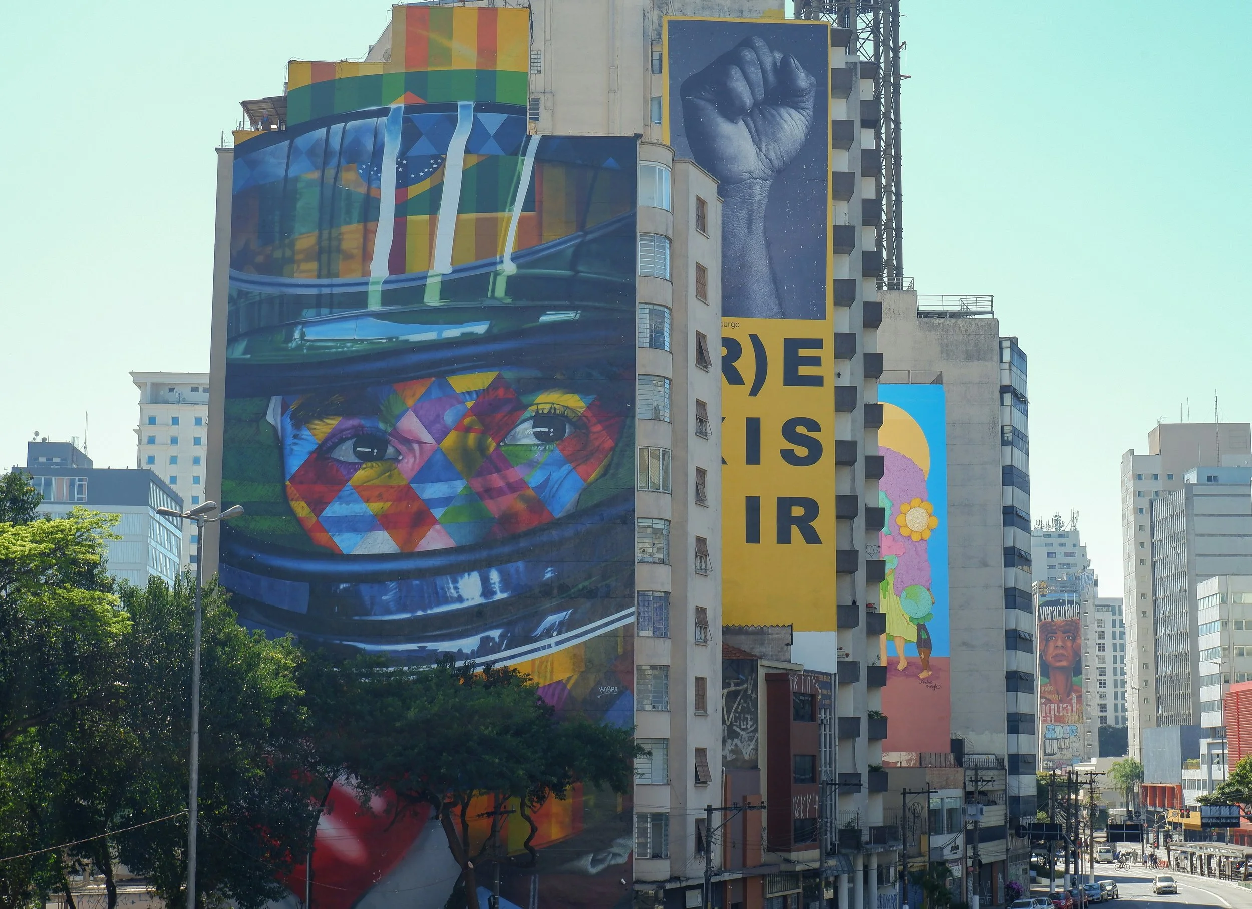 Colorful murals on tall building walls in an urban area, including large eye and fist images, with trees and other buildings in the background.