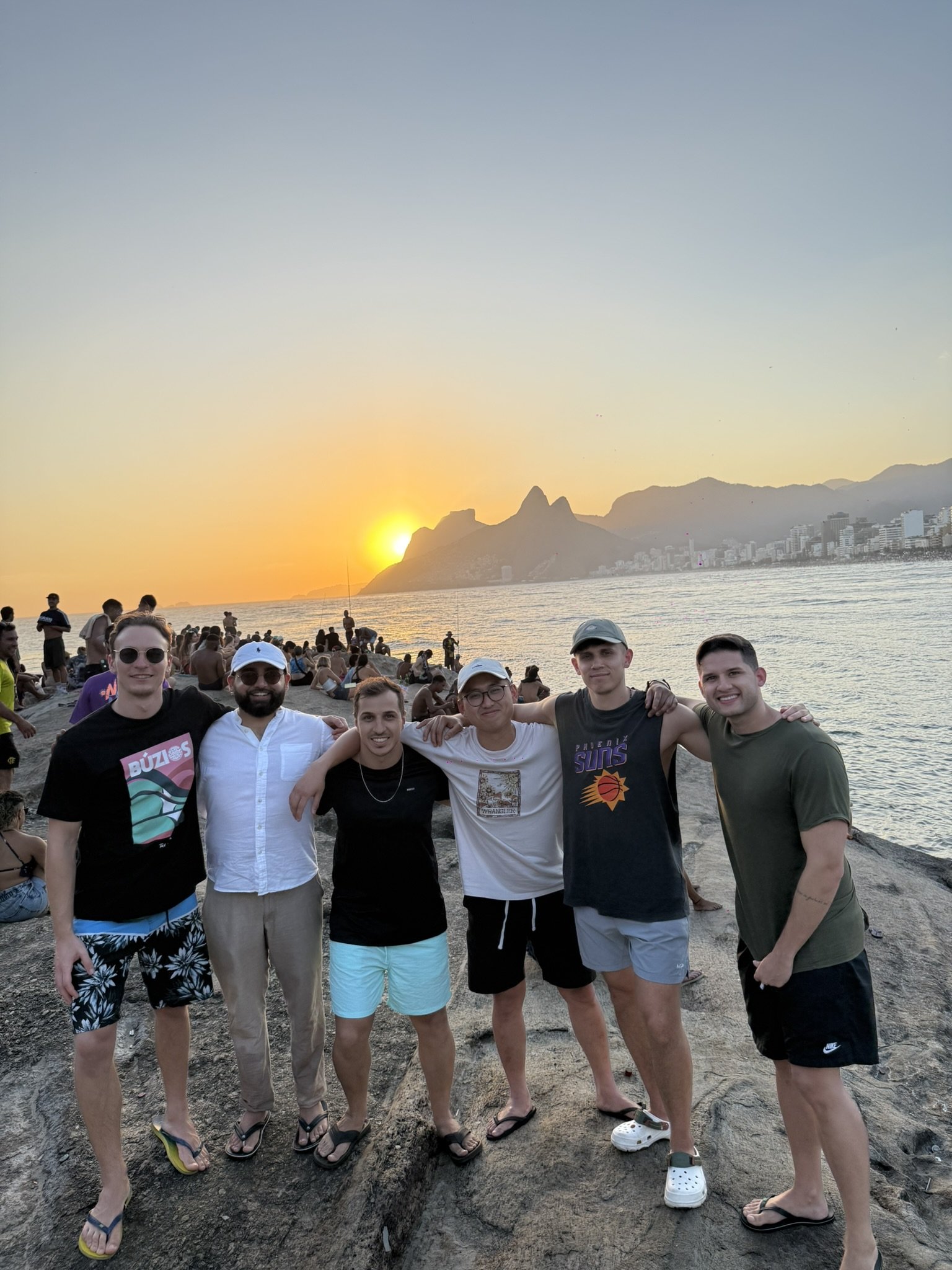 Group of young men standing on rocks at the beach during sunset, with mountains and city buildings in the background.