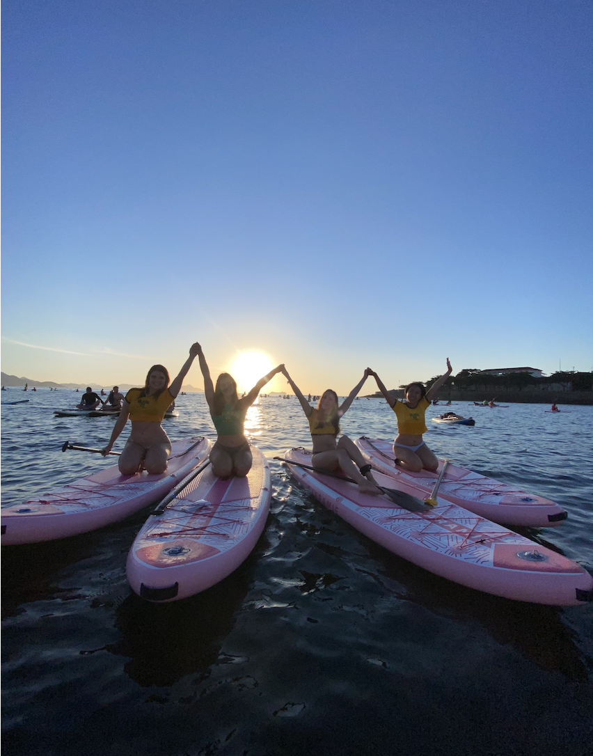 Four women kneeling on paddleboards in the water at sunset, holding hands raised in celebration.