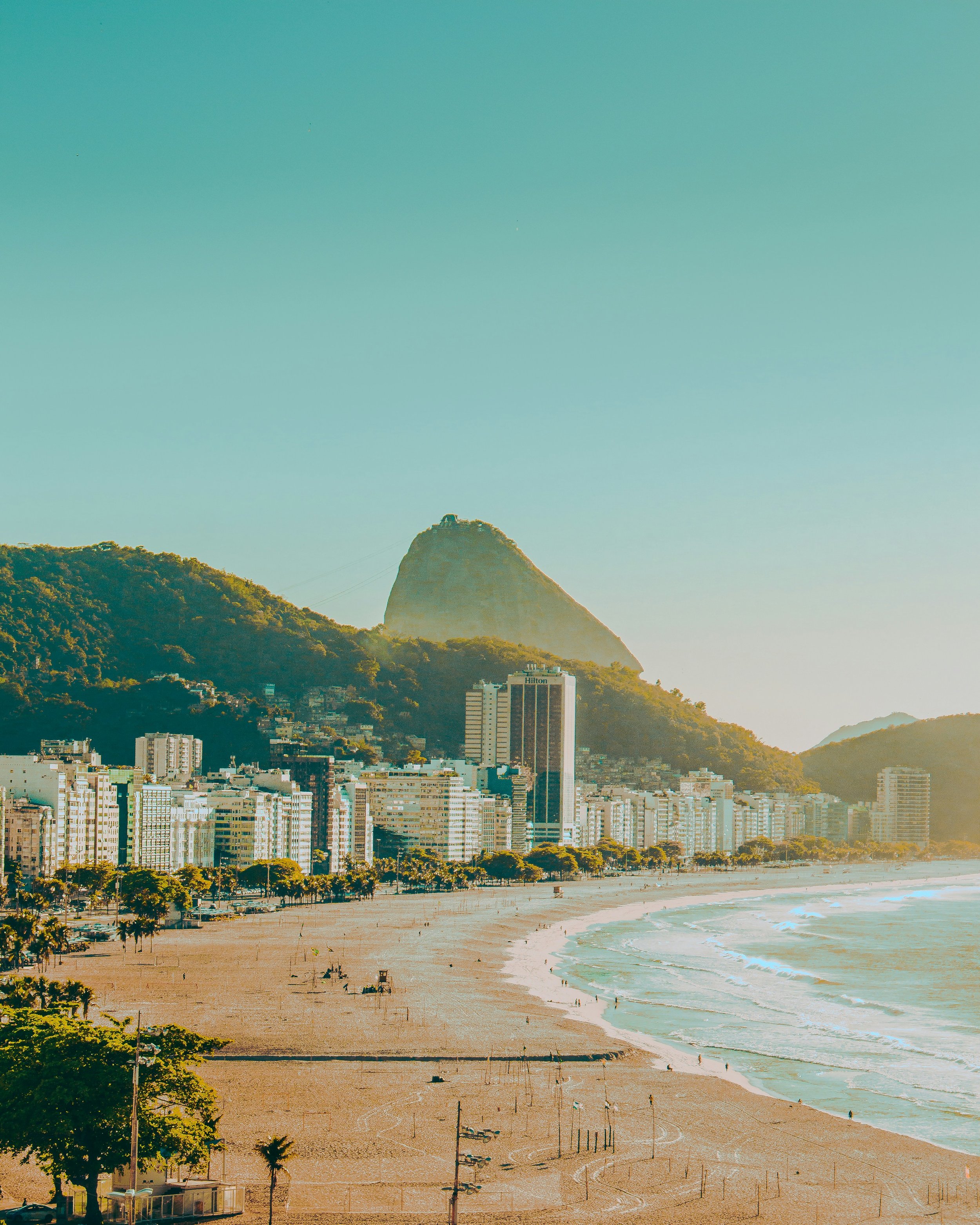 View of city skyline with tall buildings, a mountain with a cable car, and a sandy beach with people and palm trees in the foreground.