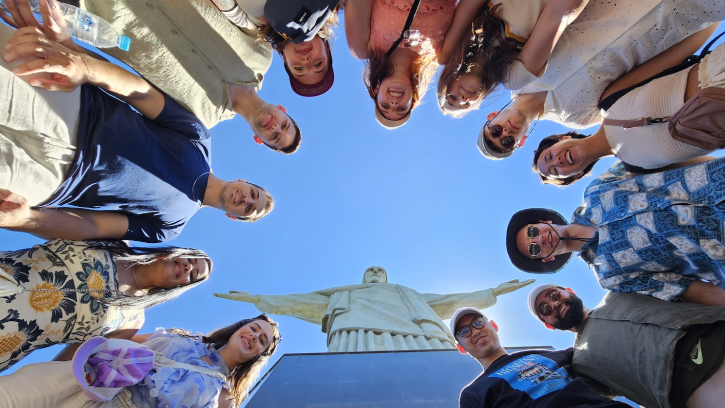 Group of people standing in a circle and looking down at the camera, with the Christ the Redeemer statue in Rio de Janeiro visible in the background, under a clear blue sky.