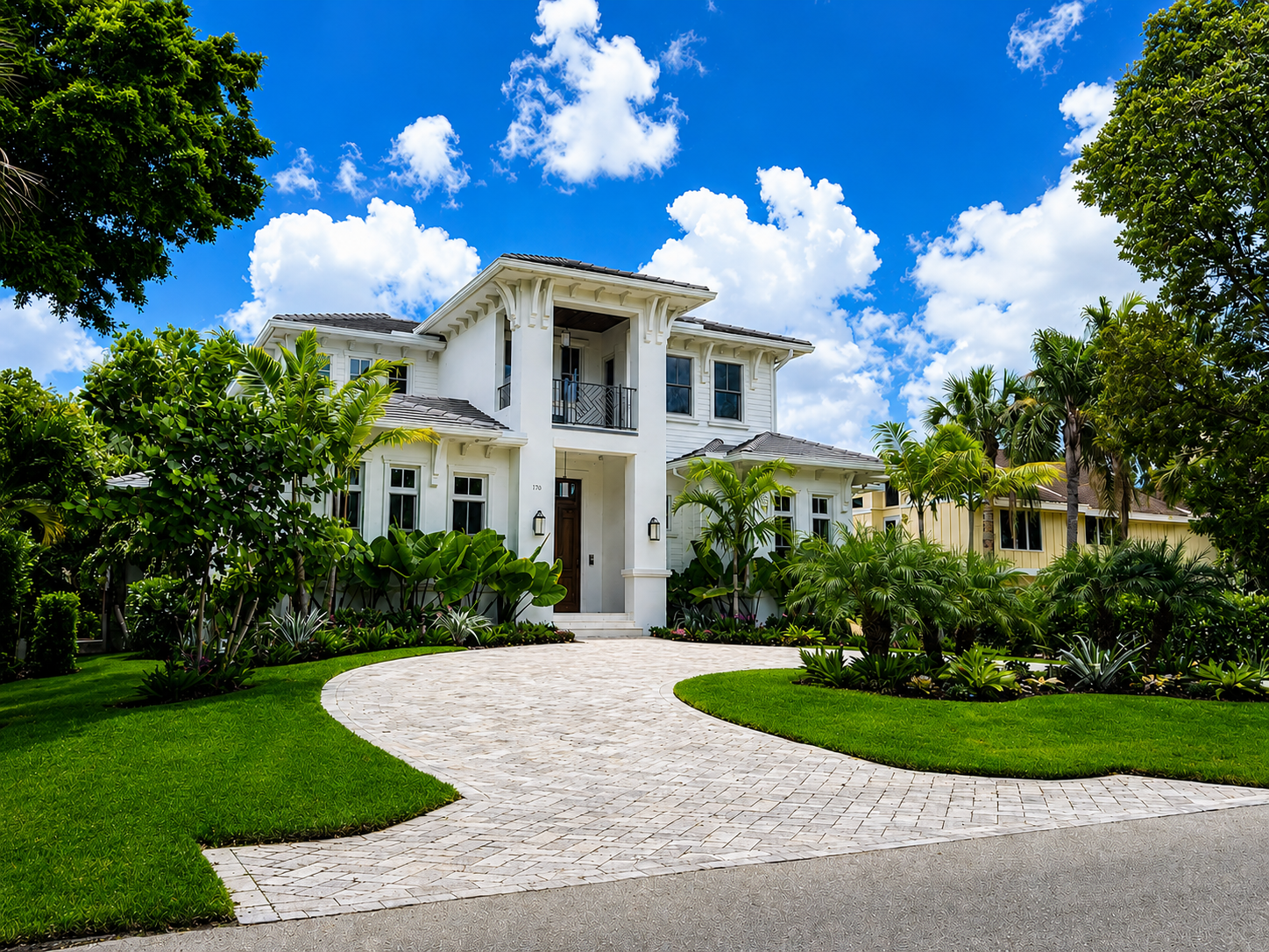A large white house with greenery and a curved stone driveway, under a bright blue sky with white clouds.