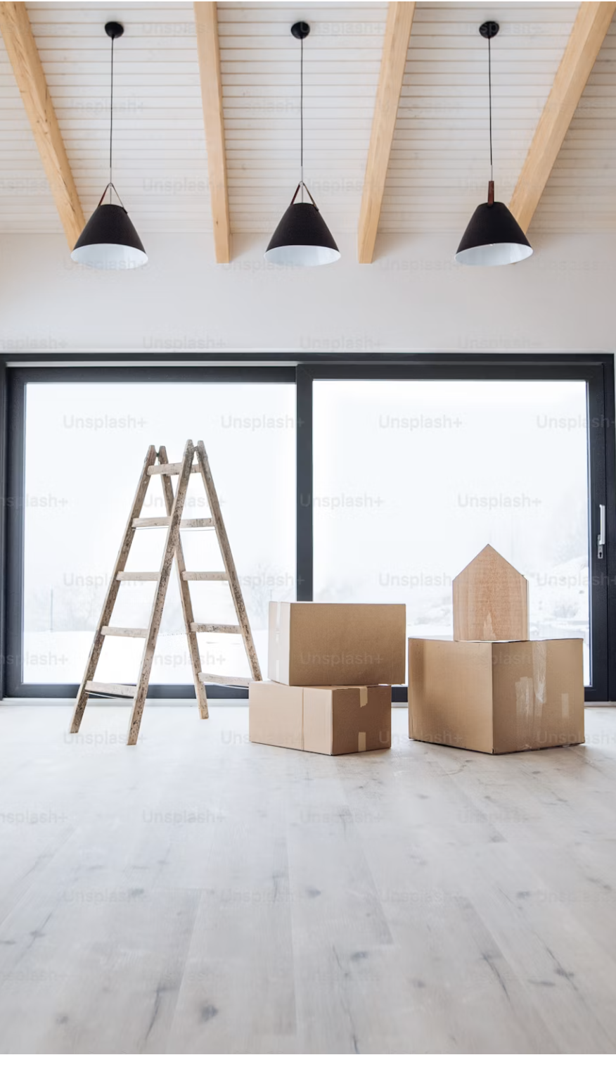 Empty room with a wooden ladder and cardboard boxes, large windows, ceiling with three hanging black pendant lights.