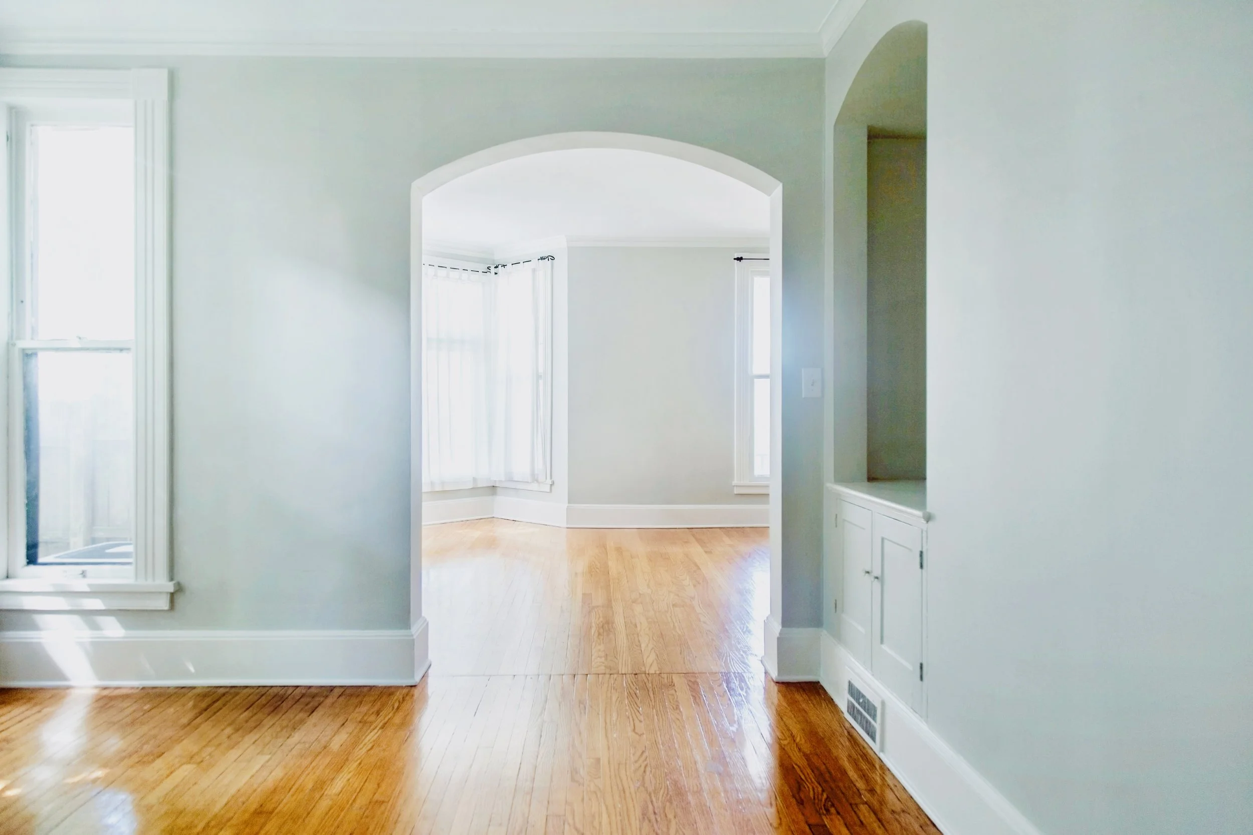 A bright, empty room with hardwood floors, white walls, large windows with white curtains, and an open archway leading to another room.