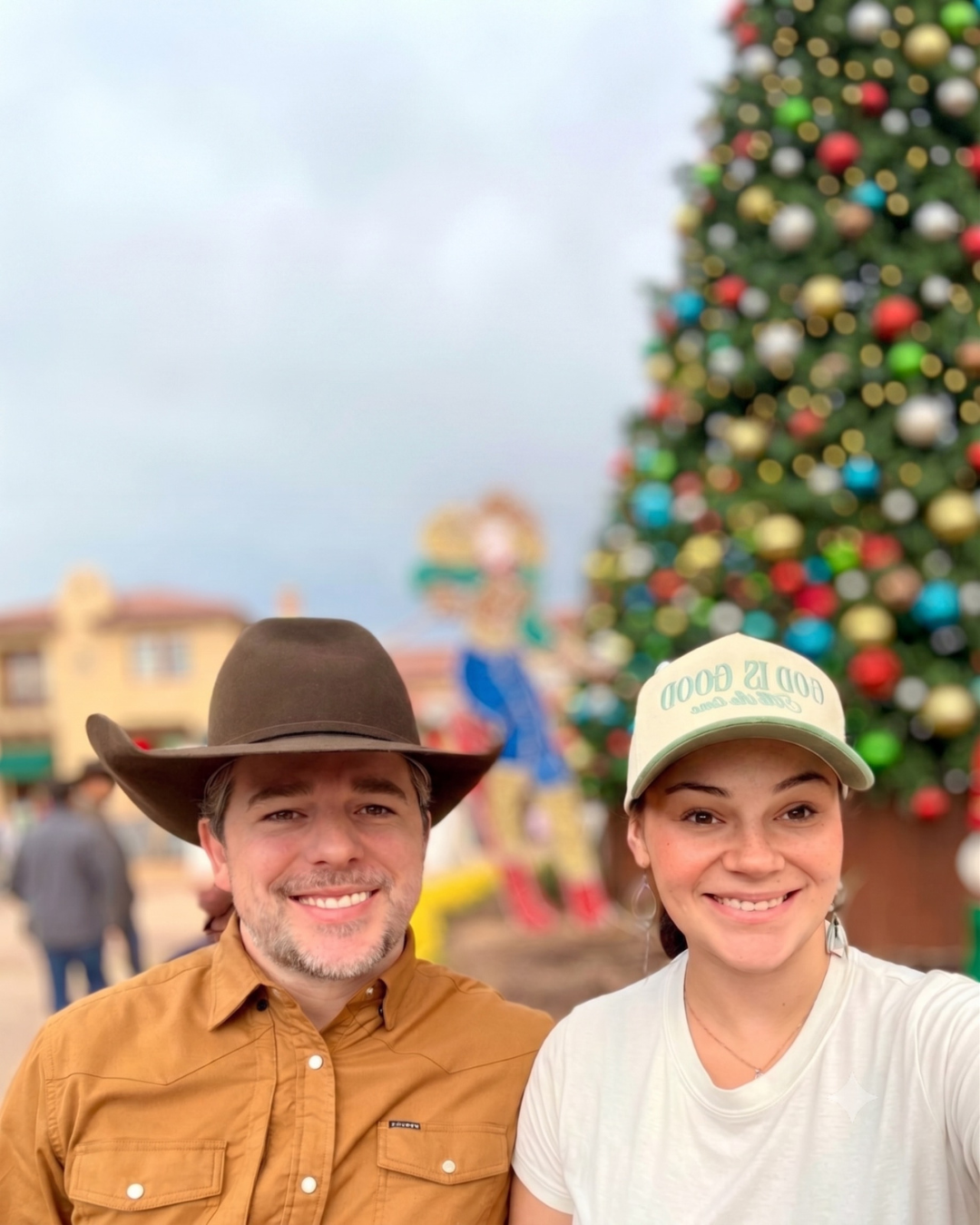 Smiling man wearing a cowboy hat and a woman wearing a 'God Is Good' cap taking a selfie in front of a decorated Christmas tree outdoors.
