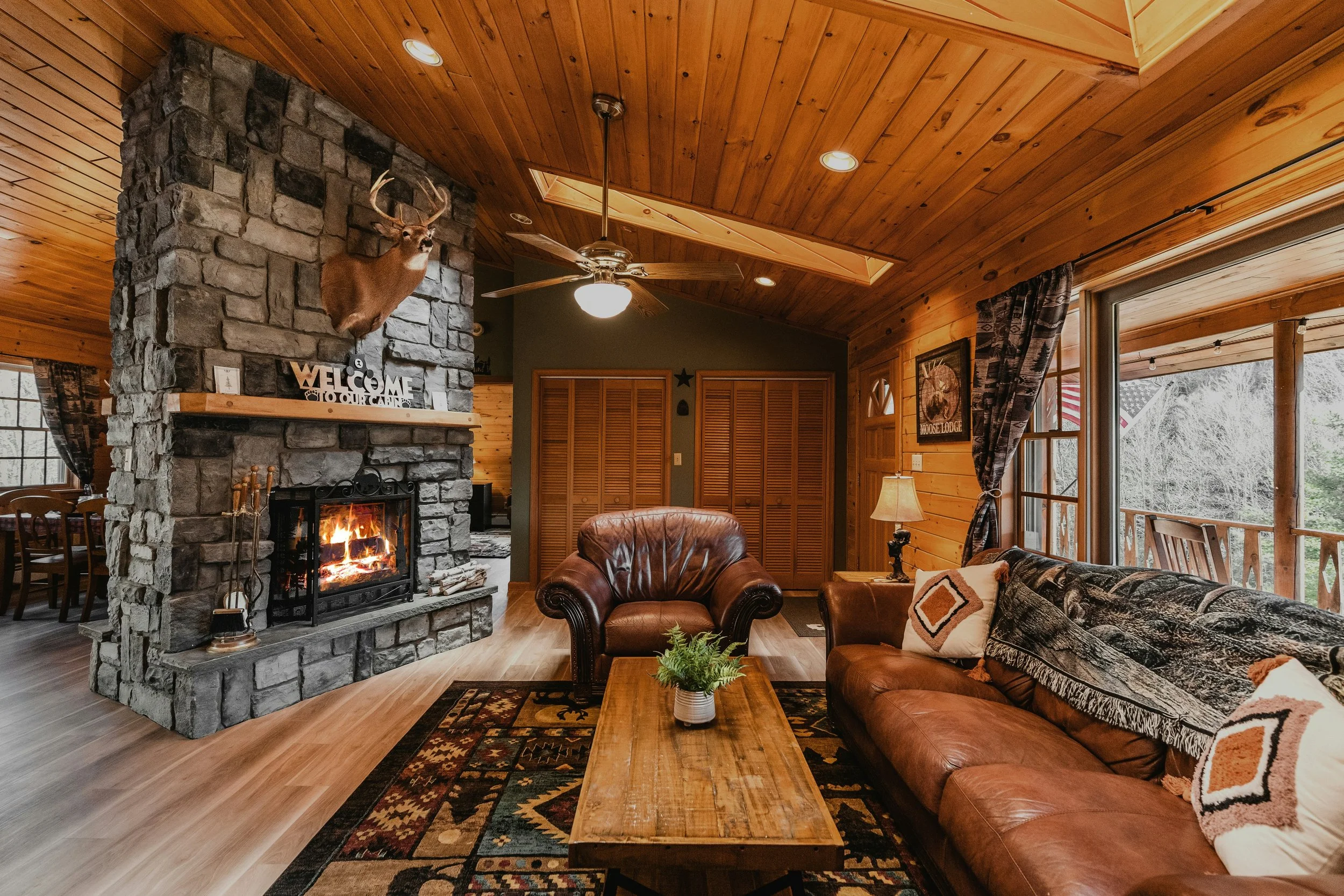 Interior of a cozy cabin living room with a stone fireplace, leather armchair, wooden coffee table, patterned rug, wood-paneled walls and ceiling, window with curtains, and a deer head mounted above the fireplace.