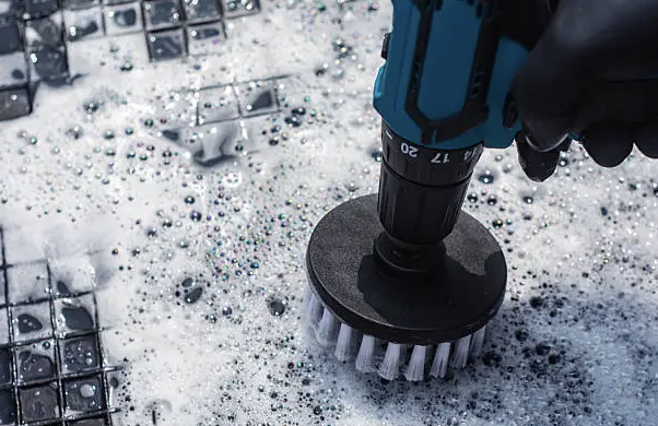 Close-up of a hand using a power drill with a brush attachment to clean a black gridded surface with soap suds and soap beads.