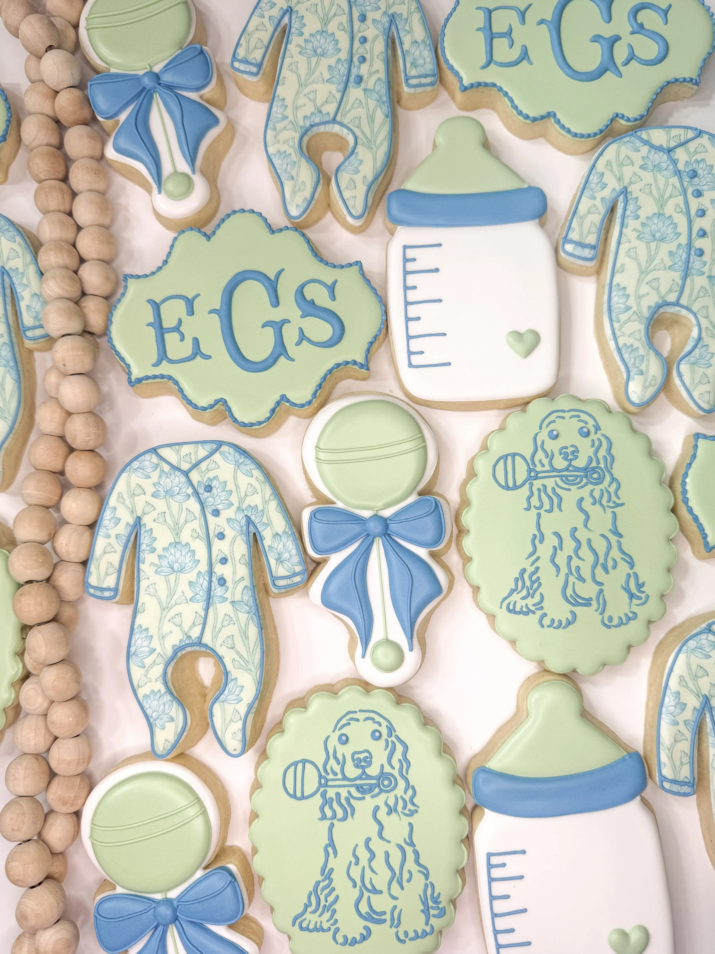 Decorated cookies with baby shower themes, including blue and green colors. Cookies shaped like baby bottles, onesies, rattles, and the word "Egs" on cloud-shaped cookies. There are also cookies with floral patterns and a string of wooden beads.