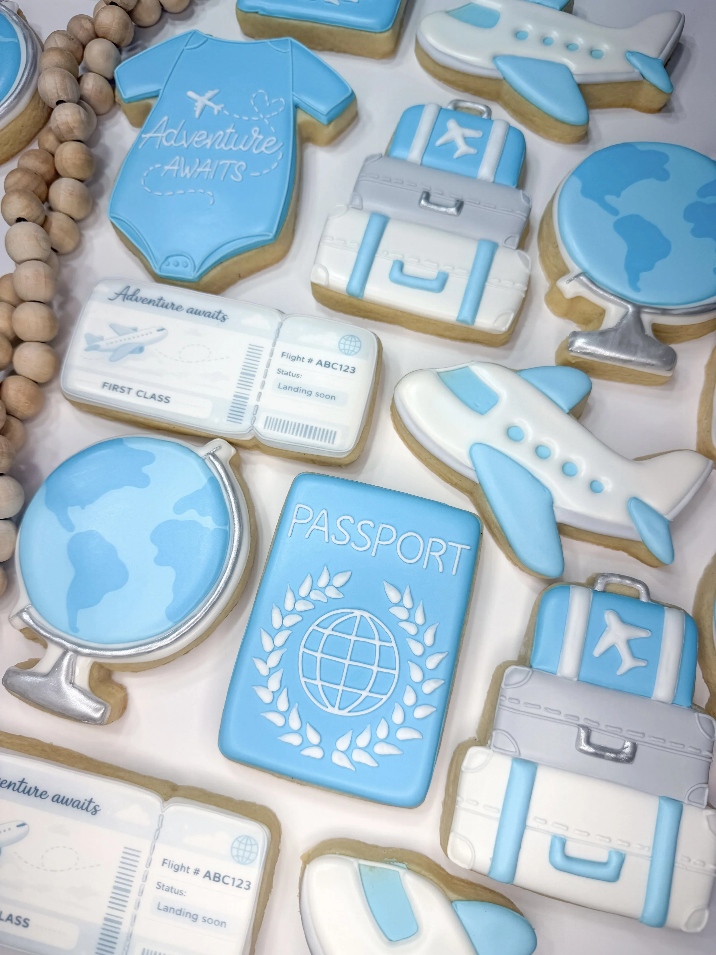 Cookies decorated with travel-themed designs including two suitcases, a globe, a passport, an airplane ticket, jet planes, and airplanes. The cookies are primarily blue, white, and silver with detailed icing.