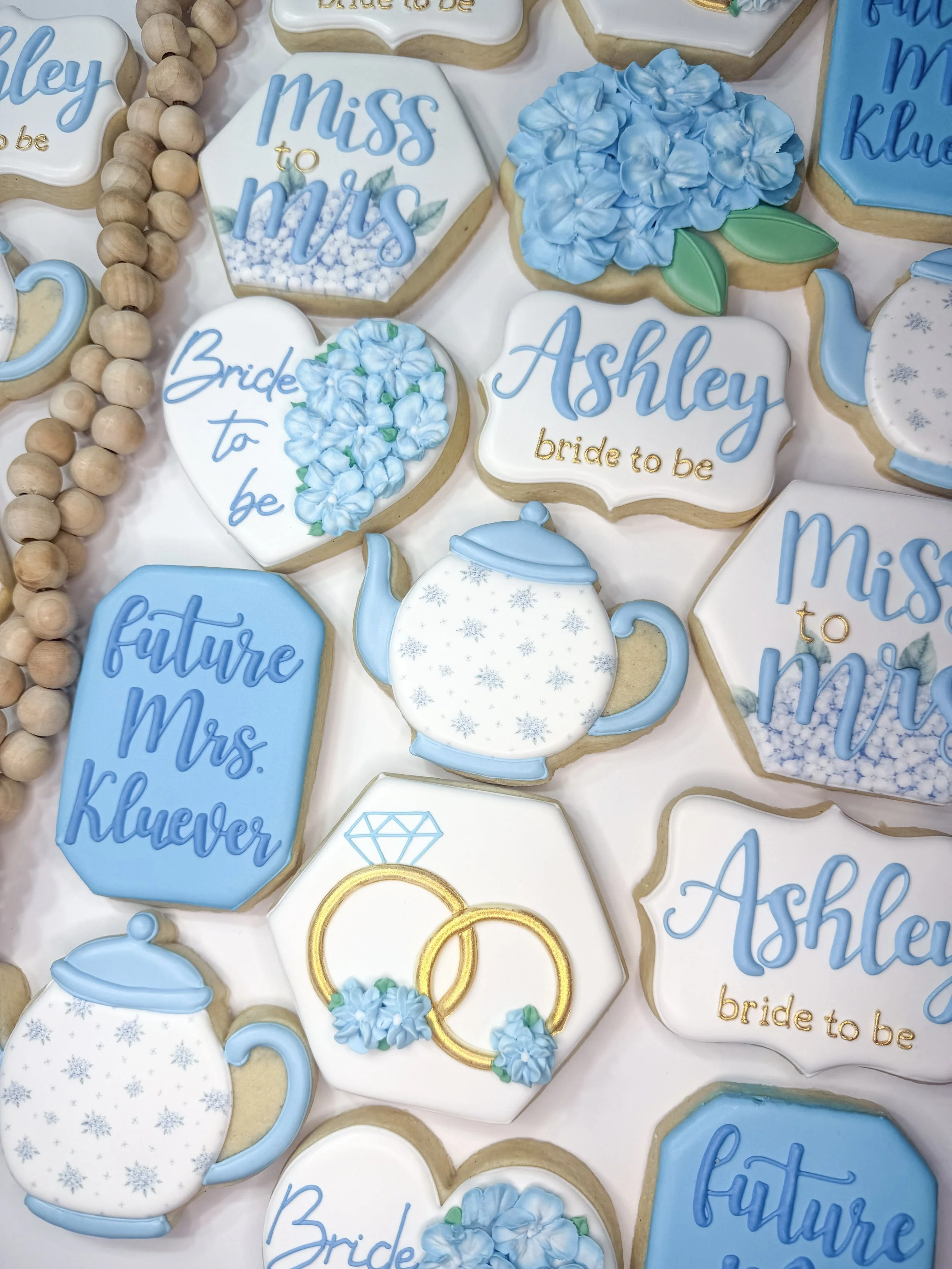 A variety of decorated cookies with blue, white, and gold icing, featuring phrases like "Miss to Mrs" and "Bride to Be," along with blue floral designs, a teapot shape, ring and diamond illustrations, and decorative beads.