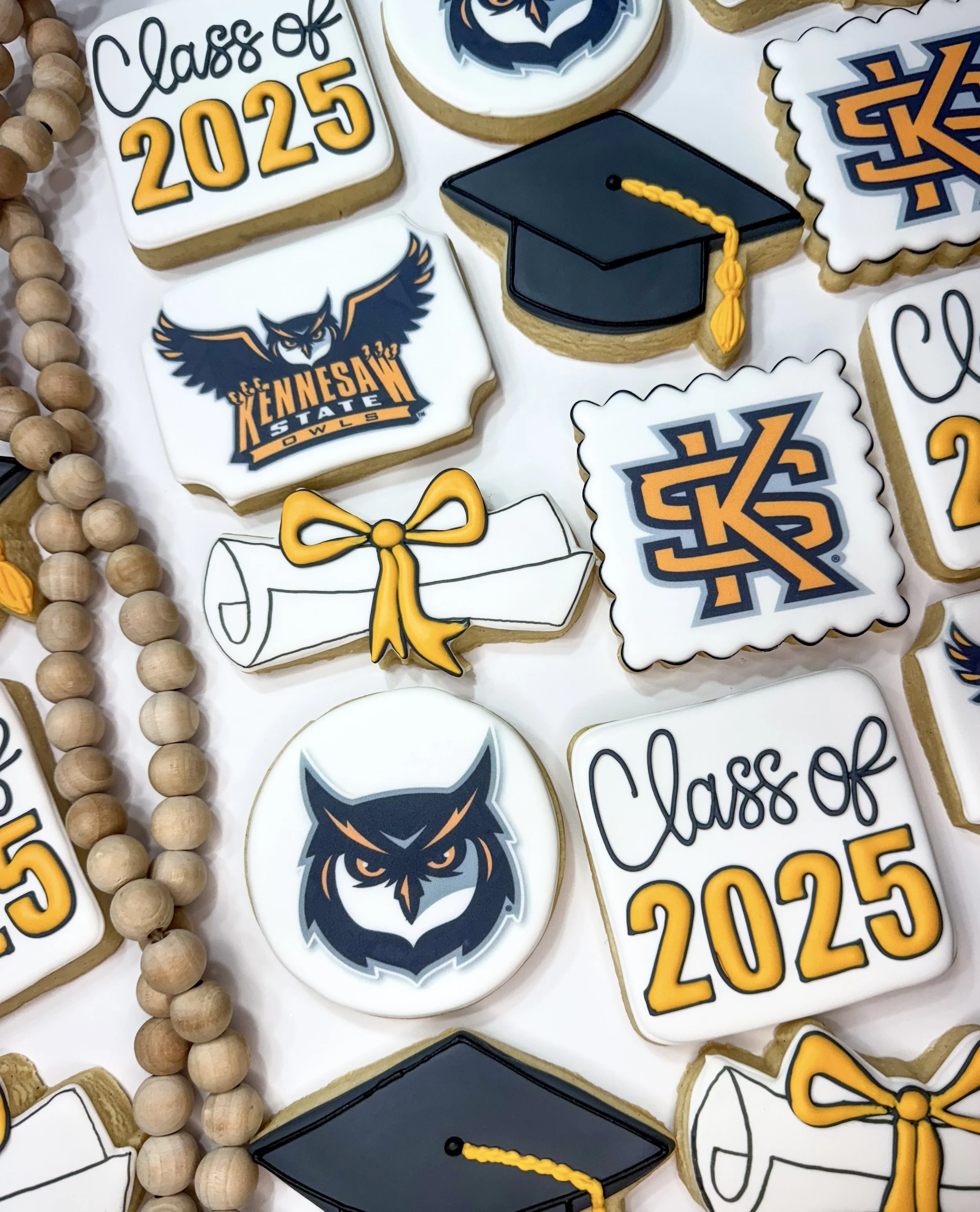 Decorated graduation cookies with the words 'Class of 2025' and the year '2025,' also featuring a college logo, a graduation cap with a yellow tassel, a bow, a diploma, and a beaded necklace.
