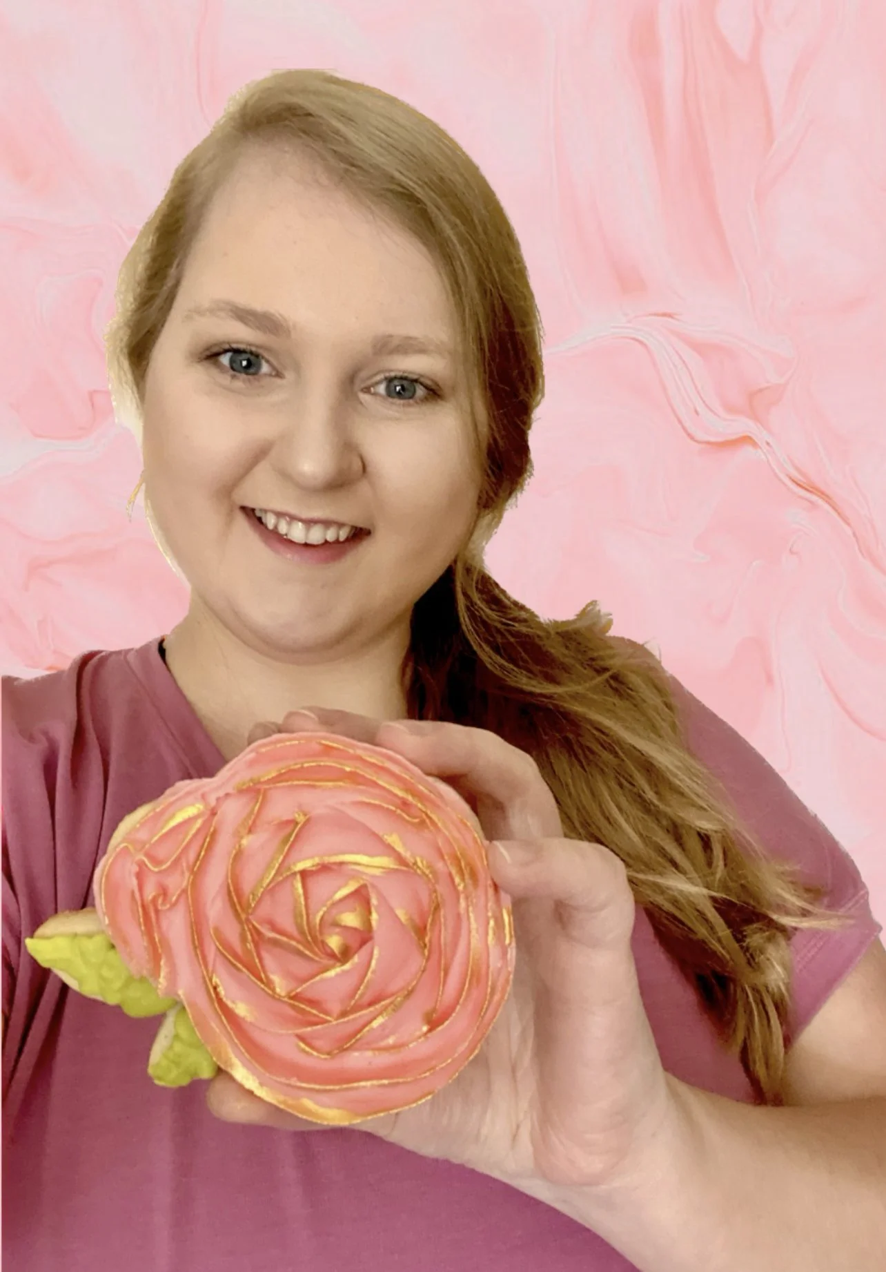 A woman with red hair and a pink shirt holds a decorated pink cookie resembling a rose, with a pink marbled background.