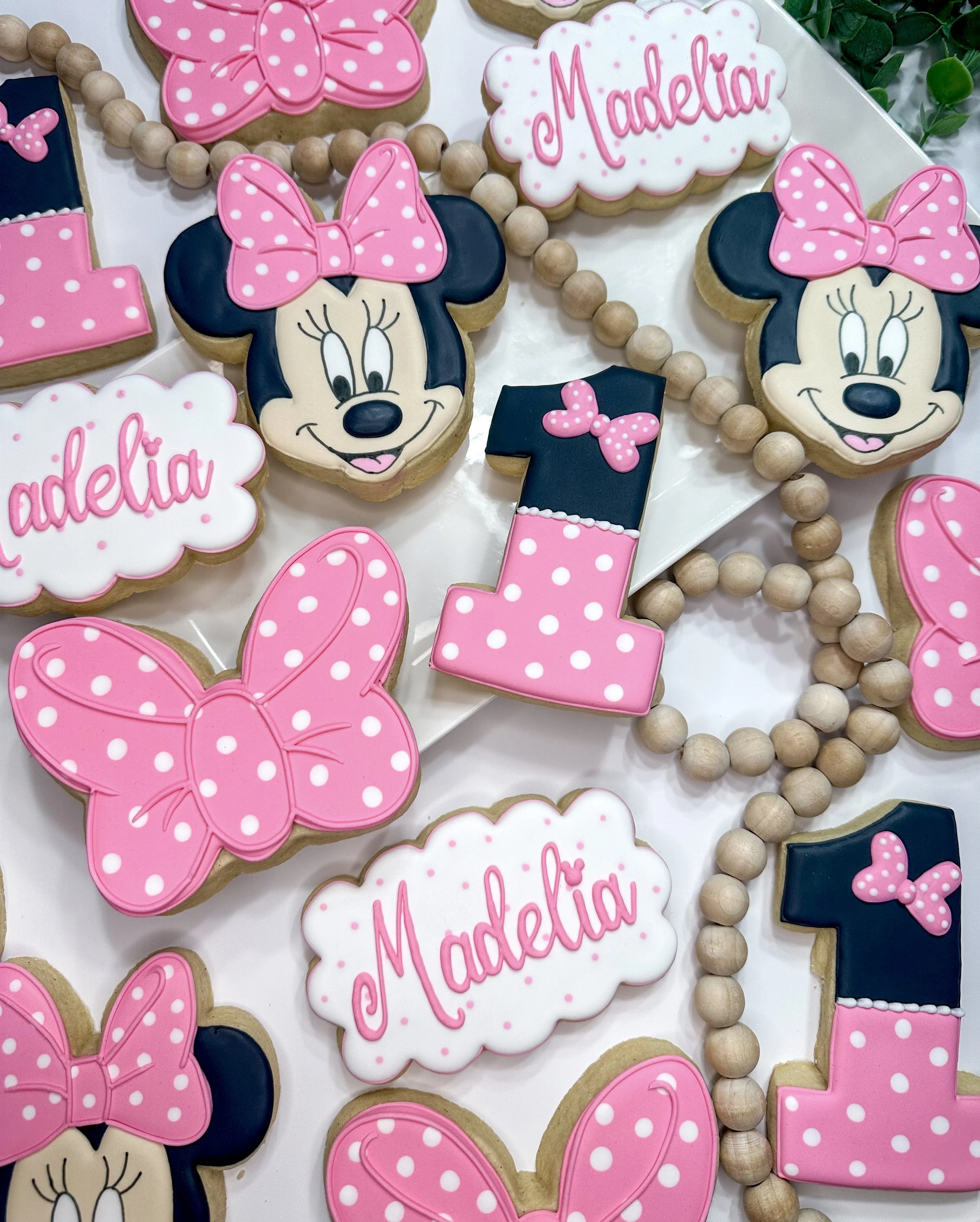 Decorated cookies with Minnie Mouse theme, pink bows, polka dots, and the name Madelia. There are also pink and white polka dot bows and the number one cookie, along with wooden beads as decoration.