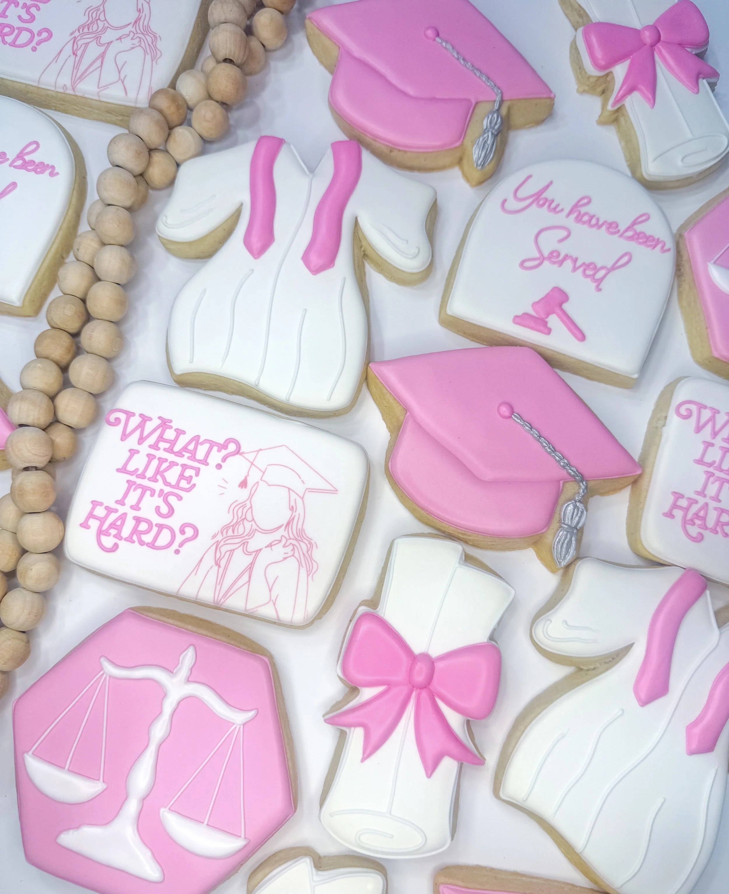 Collection of decorated cookies with pink and white icing featuring graduation-themed designs, pink bows, and phrases like "You have been served" and "What? Like it's hard?" on a white background. Wooden bead garland also visible.
