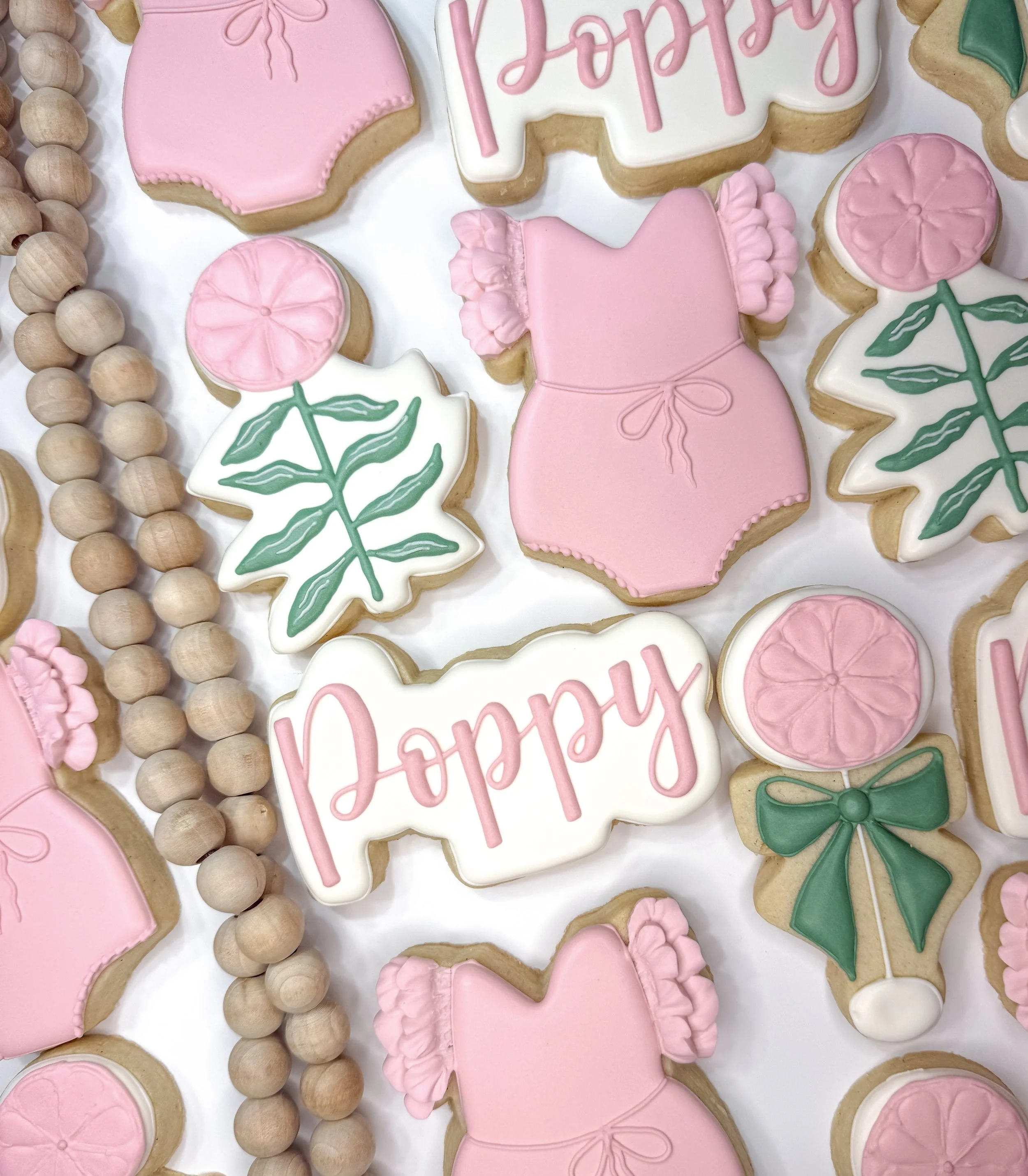 Assorted Valentine's Day cookies decorated with pink and white icing, including dresses, roses, and the word "Love" written in pink icing, surrounded by a string of wooden beads.