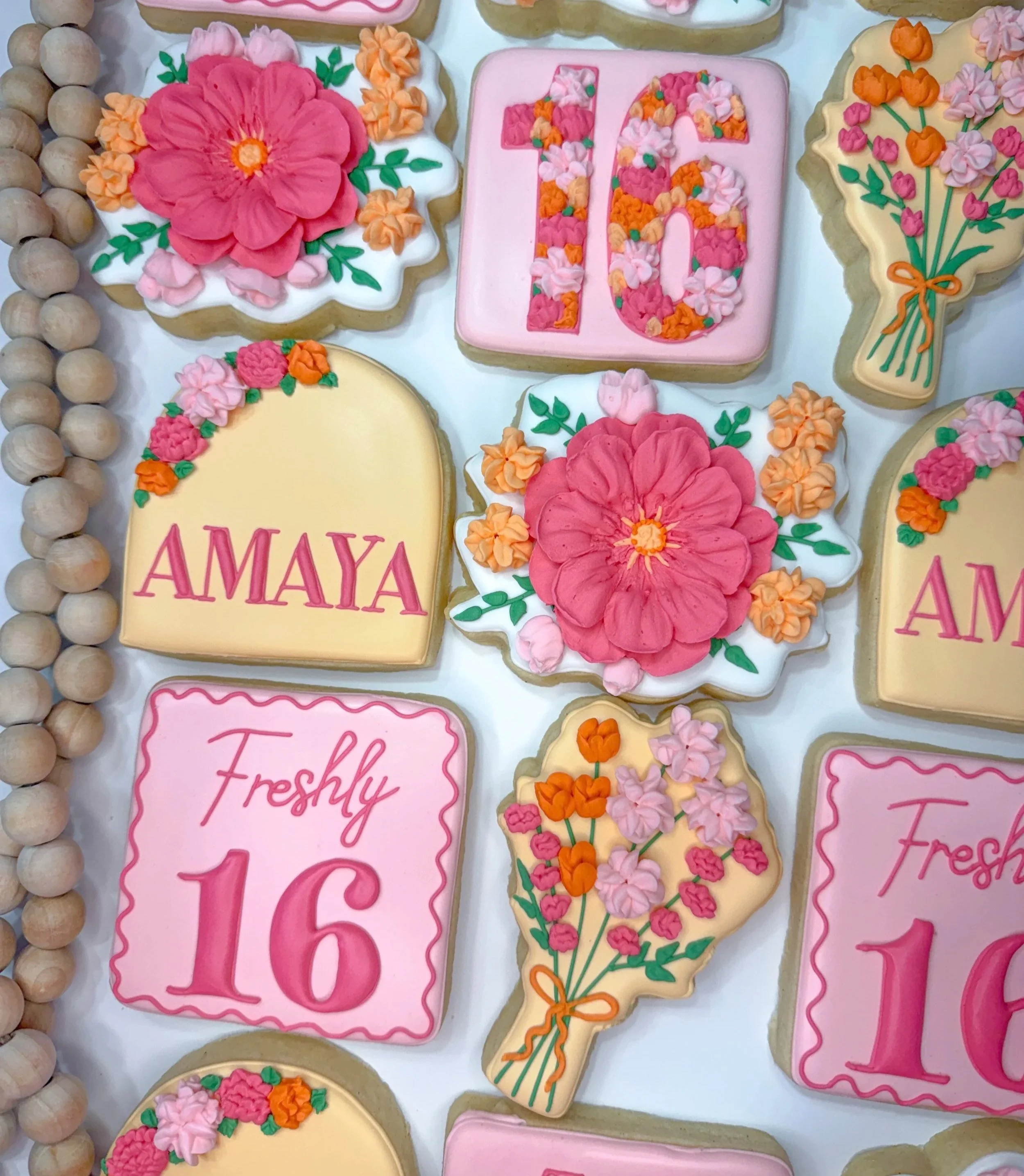 Decorated cookies with pink and peach floral icing and edible flowers, one cookie with initials 'HG' and 'AMAYA' written on it, some cookies shaped like bouquets, arranged on a white surface.