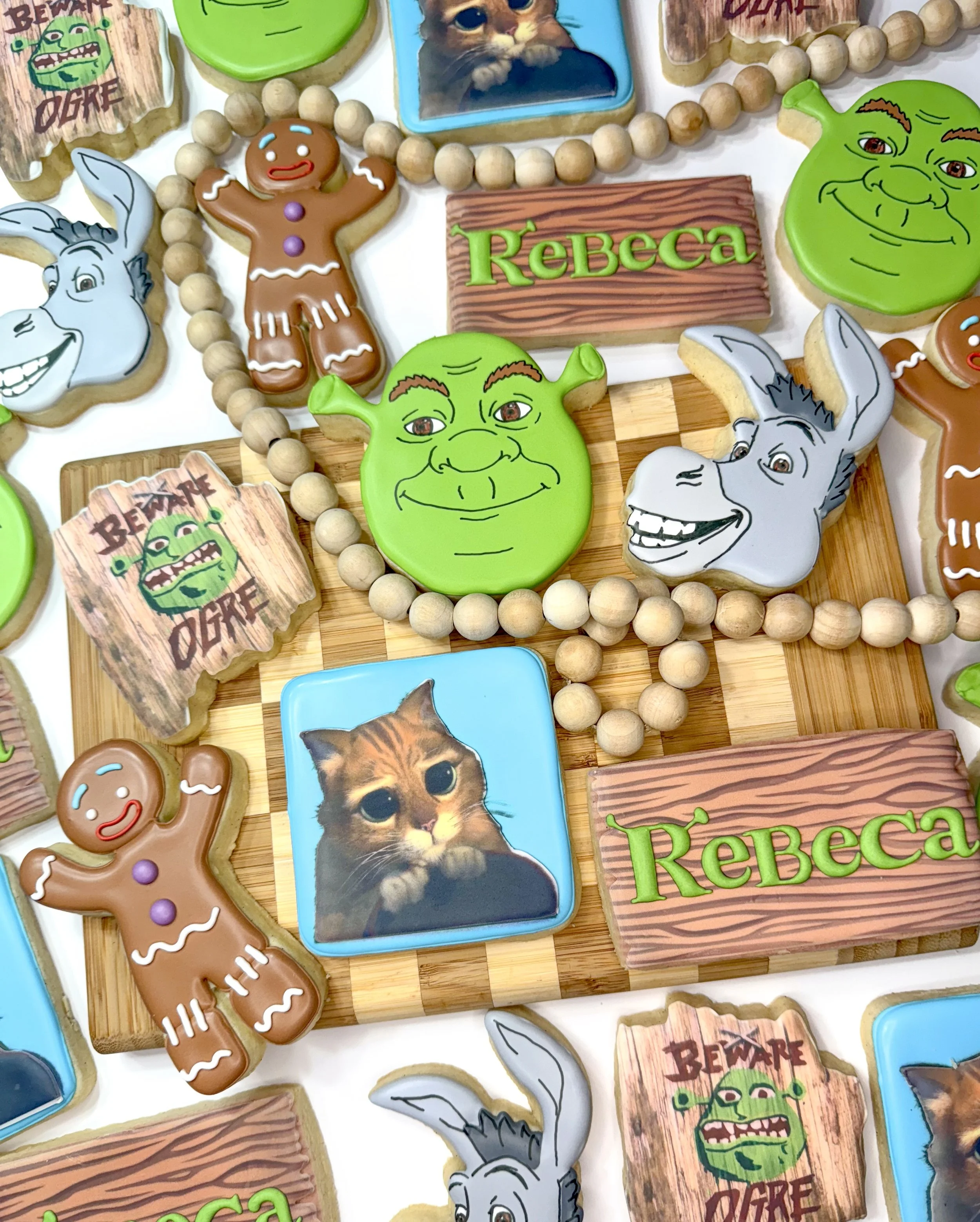 A collection of cookies decorated with characters from Shrek and others, including a gingerbread man, Donkey, Shrek, and a cat, arranged on wooden surfaces with some beaded decorations.