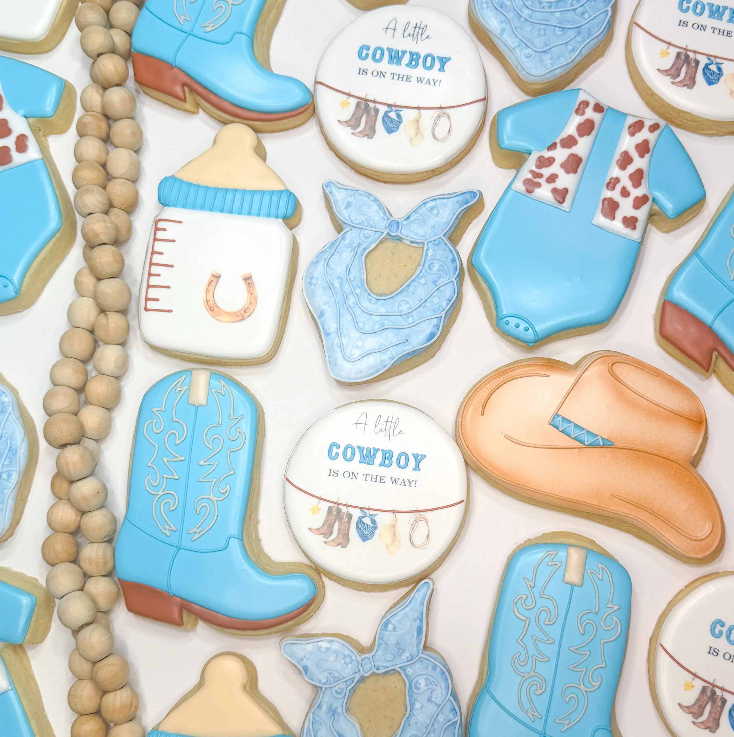 Assorted cowboy-themed cookies decorated with blue, white, and tan icing featuring cowboy boots, hats, bandanas, horseshoes, and the phrase "A little cowboy is on the way!" along with a string of wooden beads.