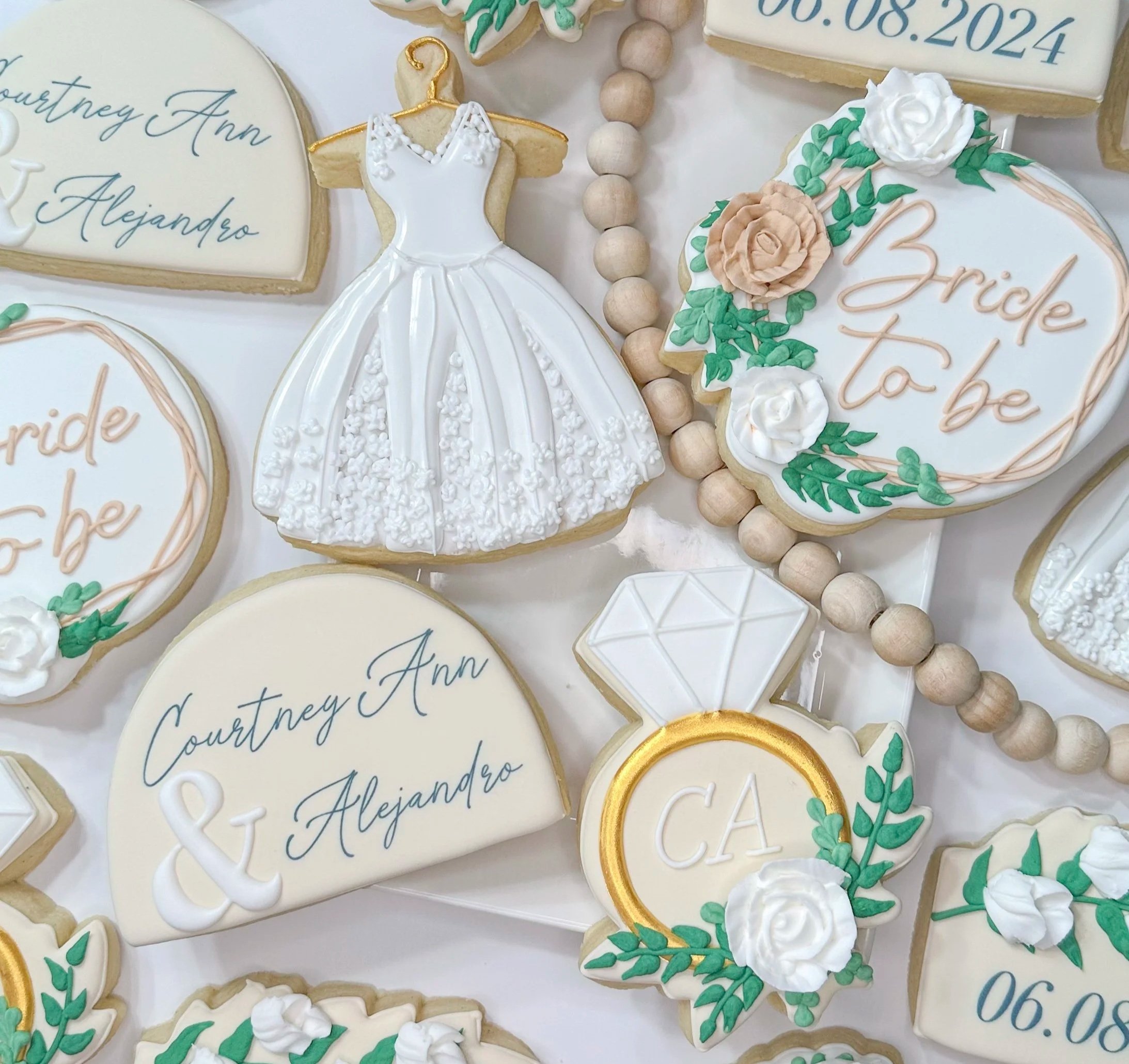 Assorted decorated cookies, including a dress-shaped cookie, heart-shaped cookies with cursive writing, and cookies with floral designs and dates, are arranged on a white surface, connected by a string of wooden beads.