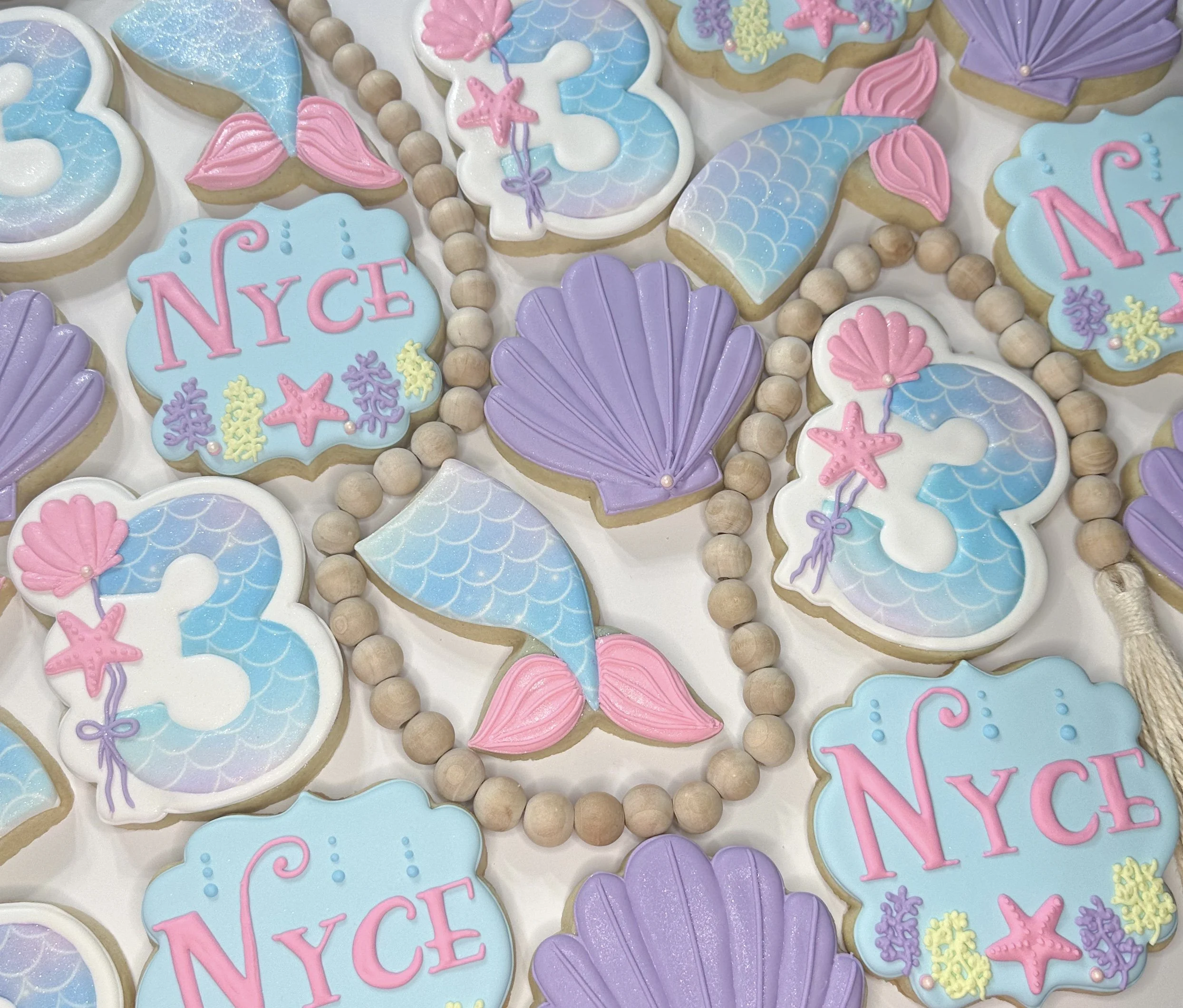 Assorted cookies decorated with ocean-themed pastel icing, featuring seashells, starfish, mermaid tails, and the pink word "NYC" on blue background.