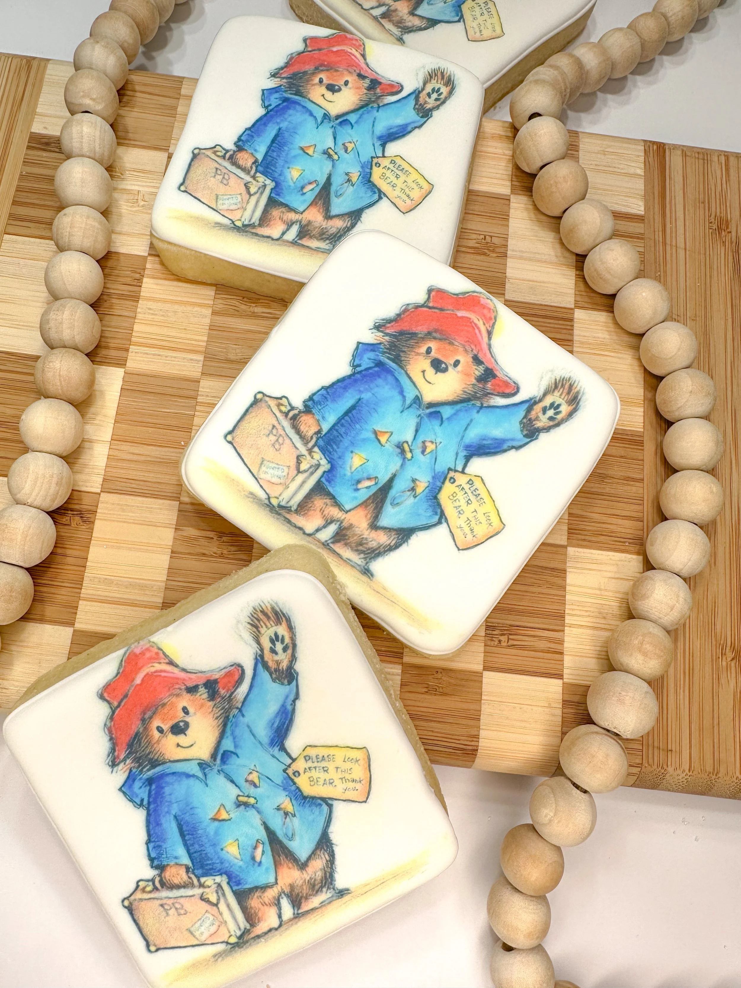 Decorative cookies featuring a bear wearing a blue coat and red hat, holding a suitcase, with a tag that says 'Please look after this bear. Thank you.'