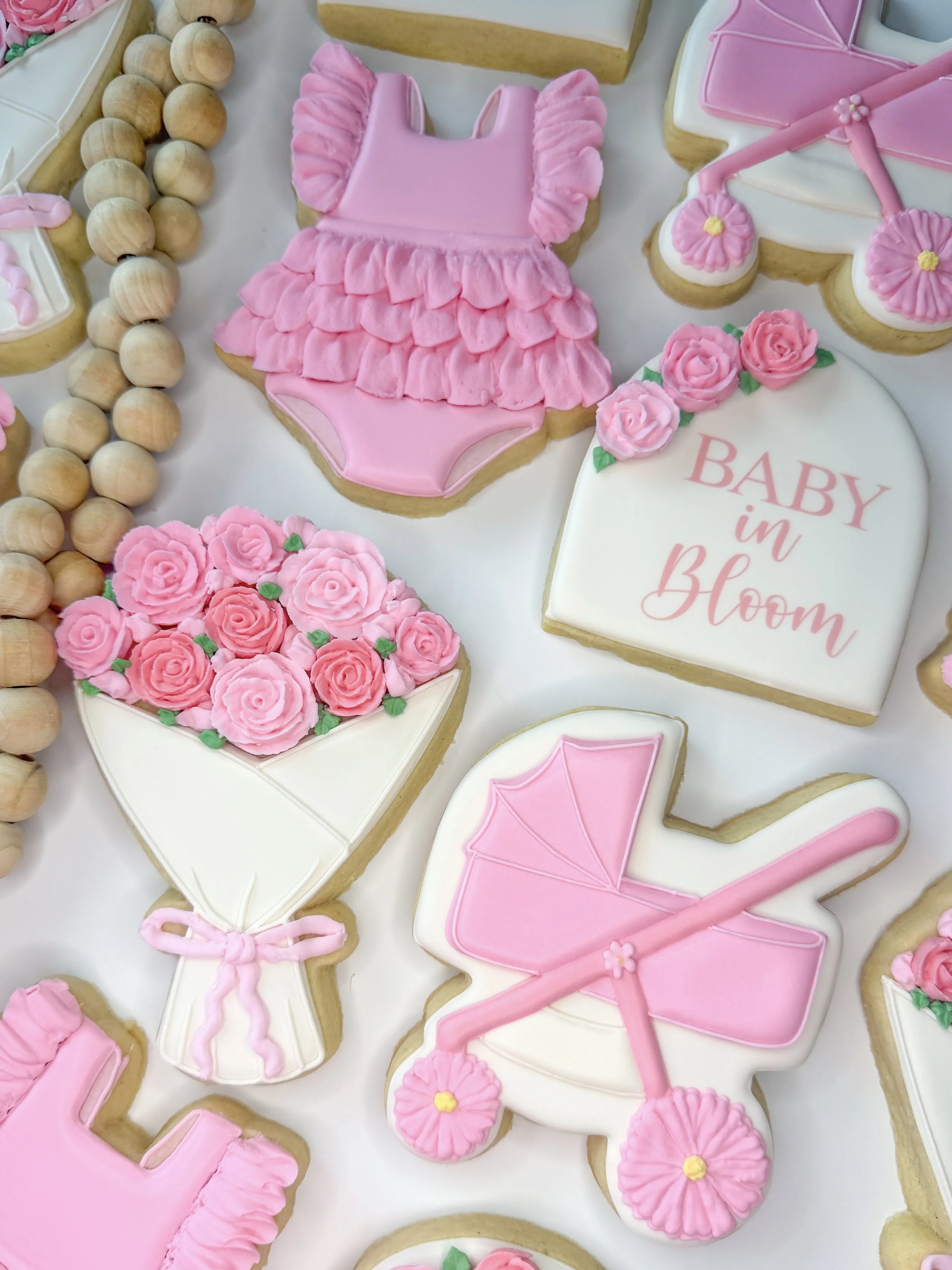 Cookies decorated with pink icing celebrating a baby shower, including a pink baby dress, pink stroller, pink flowers, and a cookie with the words "Baby in Bloom".