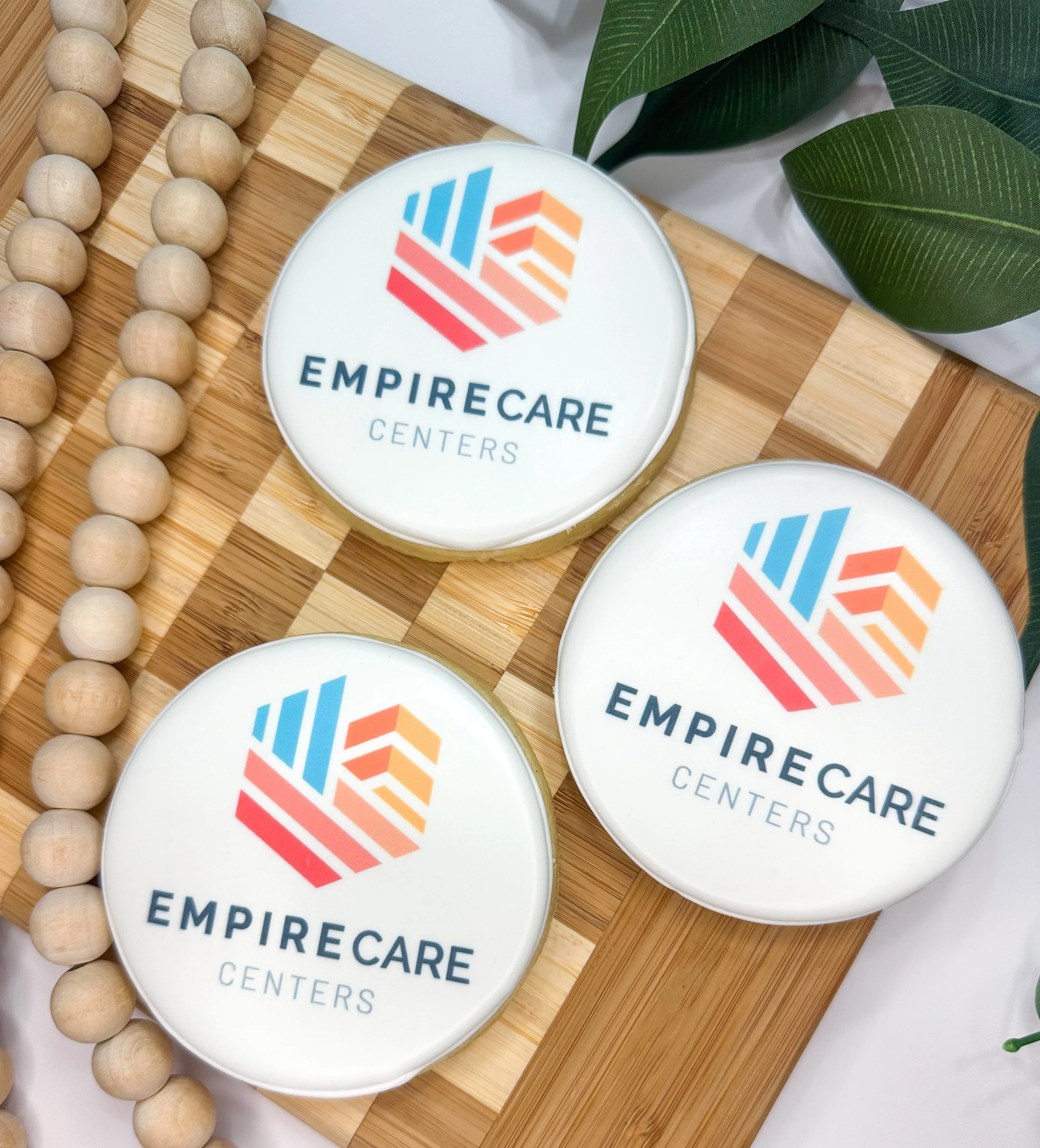Close-up of a round sign with the EmpireCare Centers logo, featuring a colorful geometric design, placed on a wooden surface next to a string of wooden beads and green leaves.