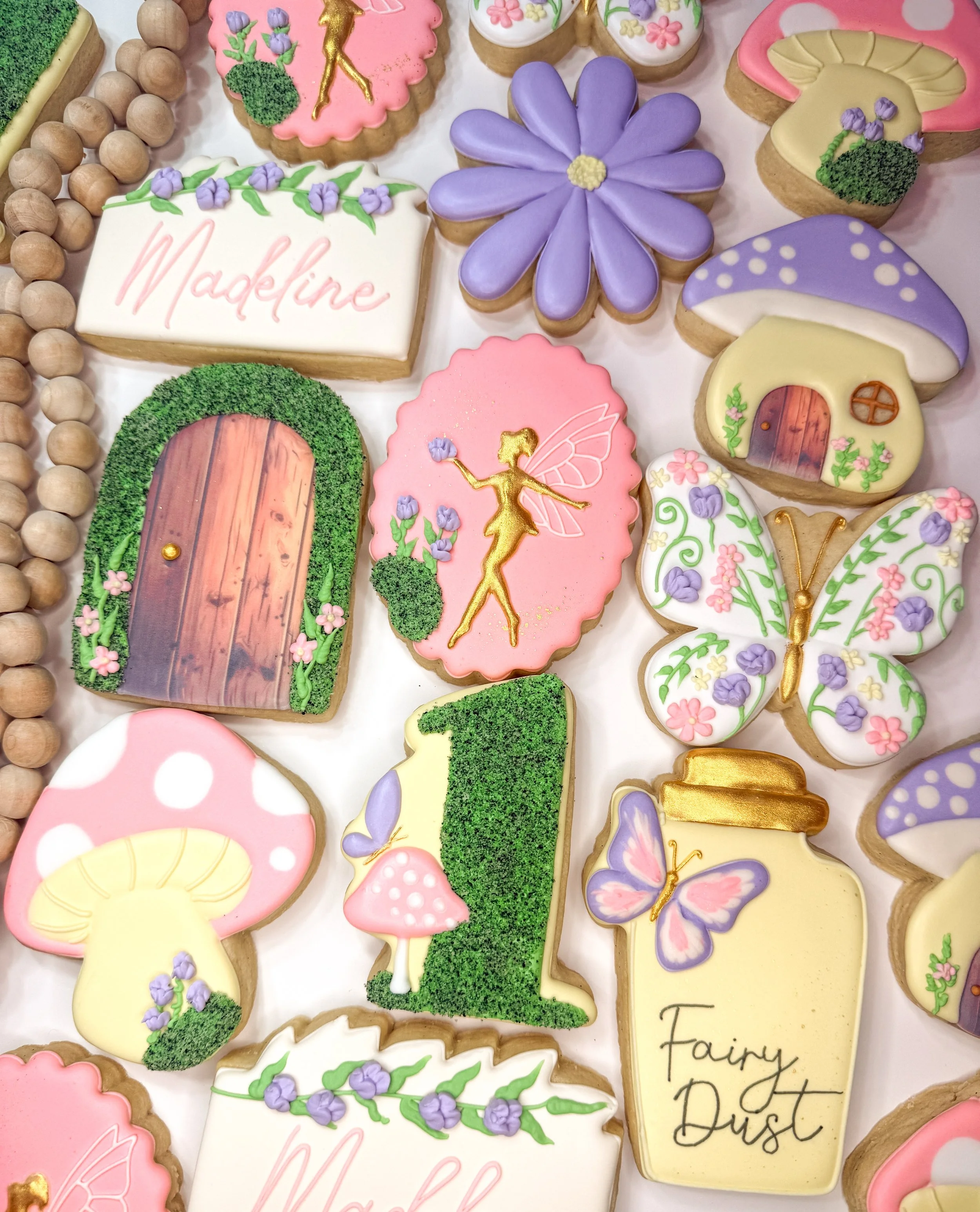 Collection of decorated cookies with fairy tale theme, including a pink cheeked cookie with a gold fairy silhouette, a large purple flower, a green door, a fairy dust label, and butterflies, some with floral patterns.