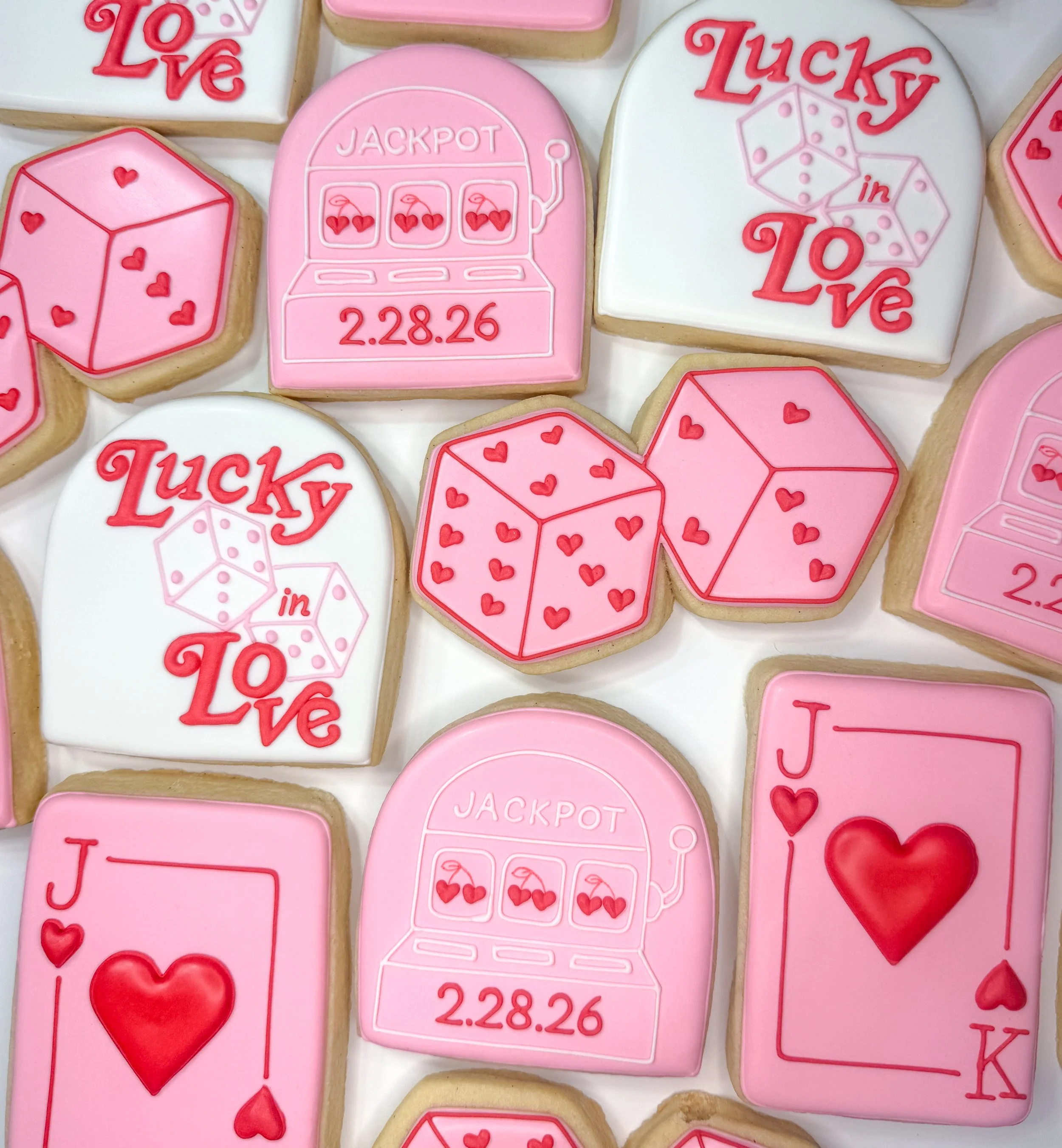 Decorated cookies with Valentine's Day love and luck themes, featuring pink, white, and red icing, including a slot machine, dice, hearts, and the words 'Love' and 'Lucky in Love.'