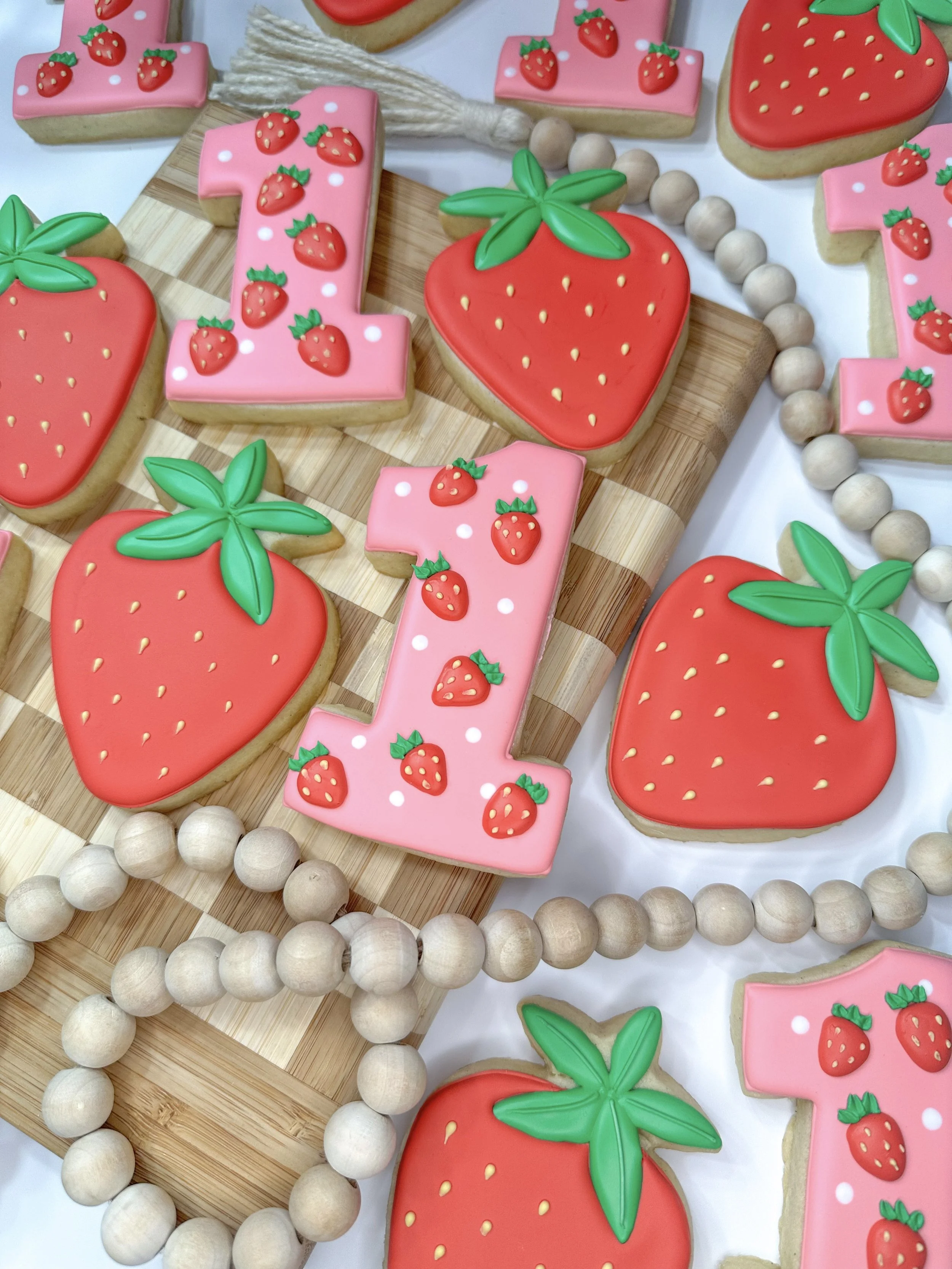 Assorted strawberry-themed decorated cookies shaped like strawberries, hearts, and the number '1', with pink and red frosting and green leaves, arranged on a wooden tray and surrounded by a string of wooden beads.