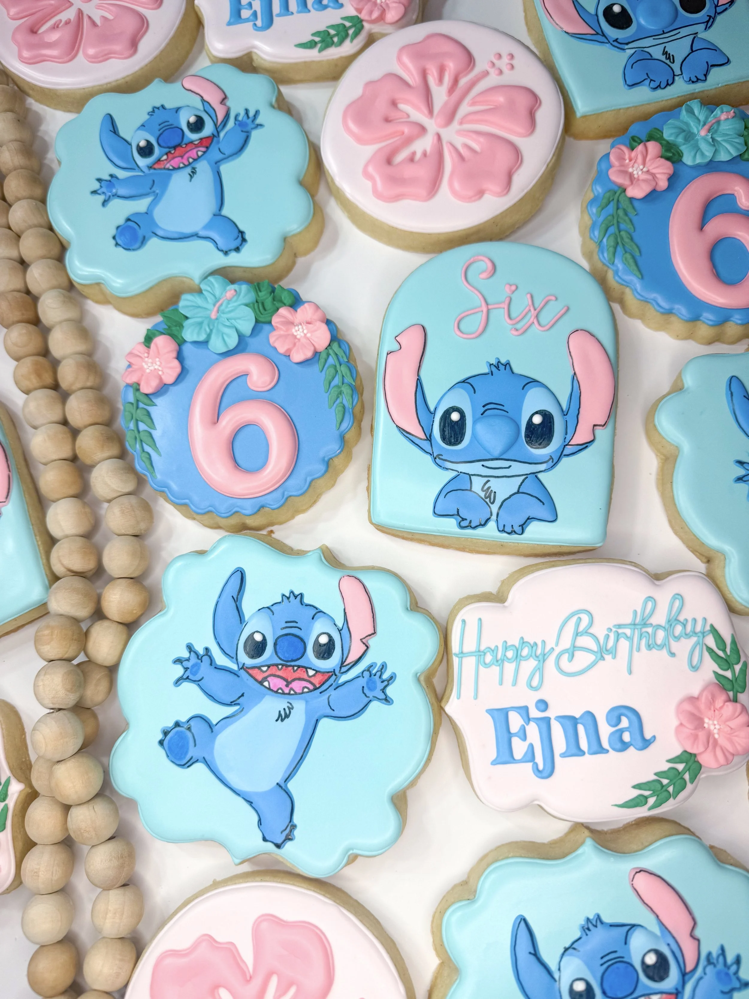 Colorful birthday cookies decorated with Stitch, flowers, and the words 'Six,' 'Happy Birthday,' and the name 'EJLA,' arranged on a white surface with a border of round wooden beads.